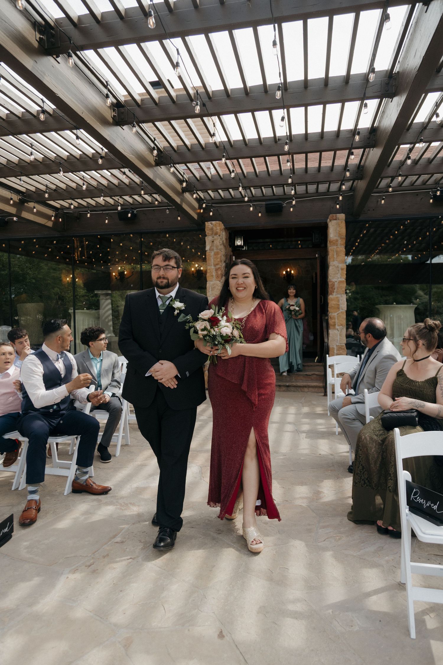 Bridesmaid in red dress walks down aisle with groomsman under pergola with string lights.