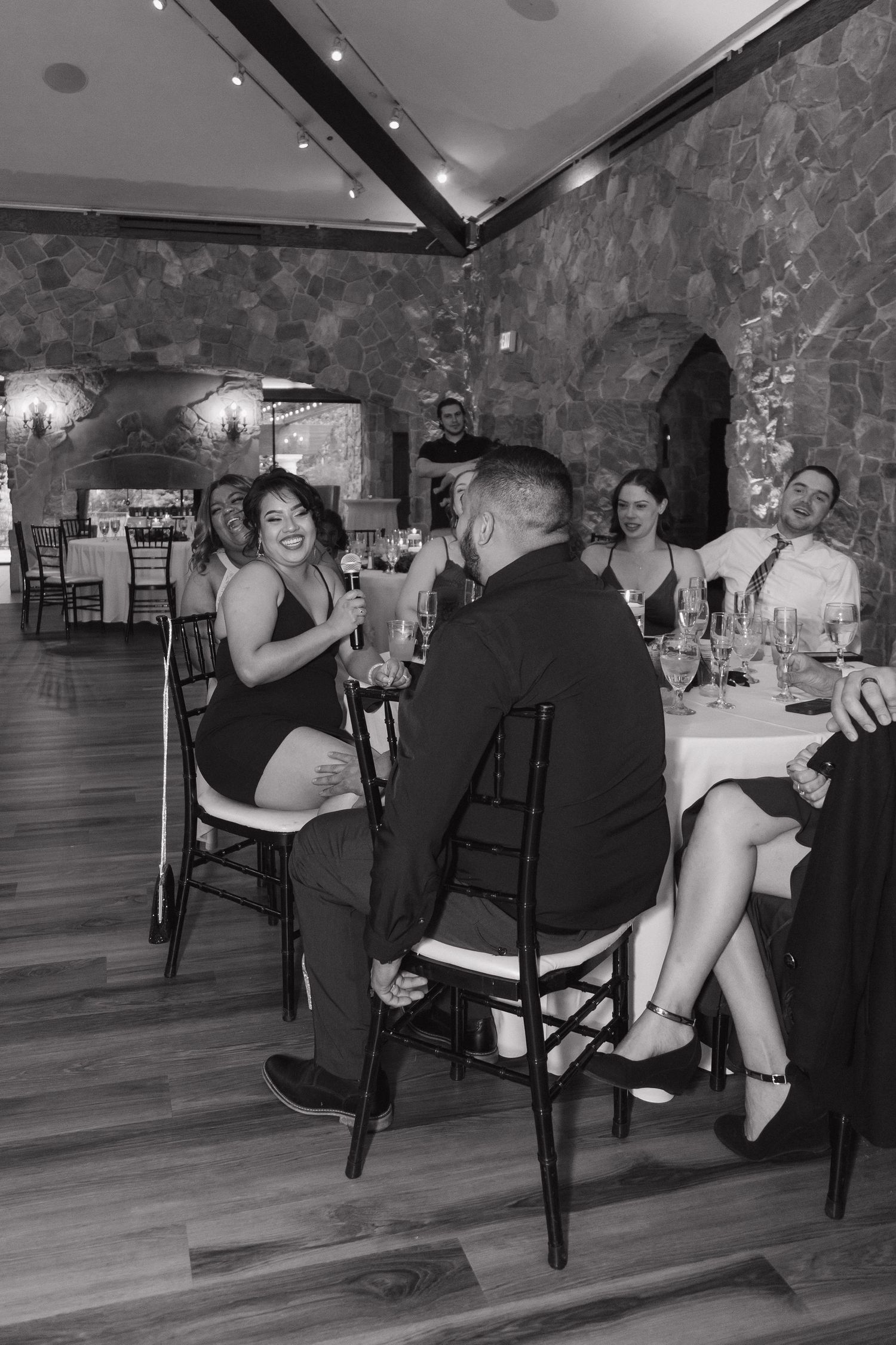 Black and white photo of guests laughing and toasting at wedding reception dinner table.