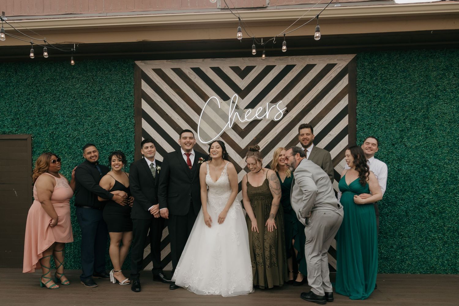 Bride and groom pose with wedding party guests in front of a green hedge wall with Cheers sign.