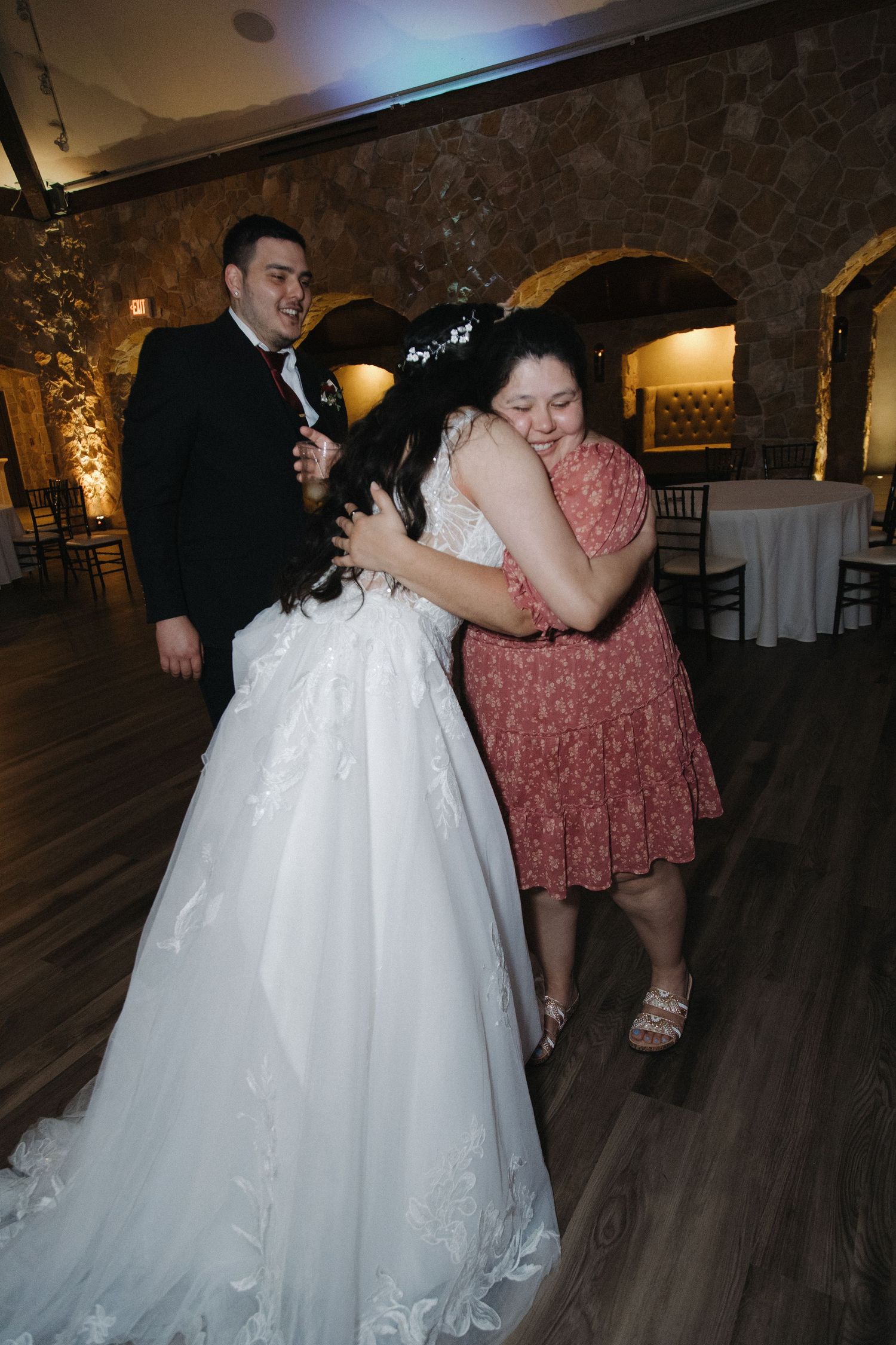 Bride in lace gown shares warm embrace with guest while groom smiles at wedding reception.