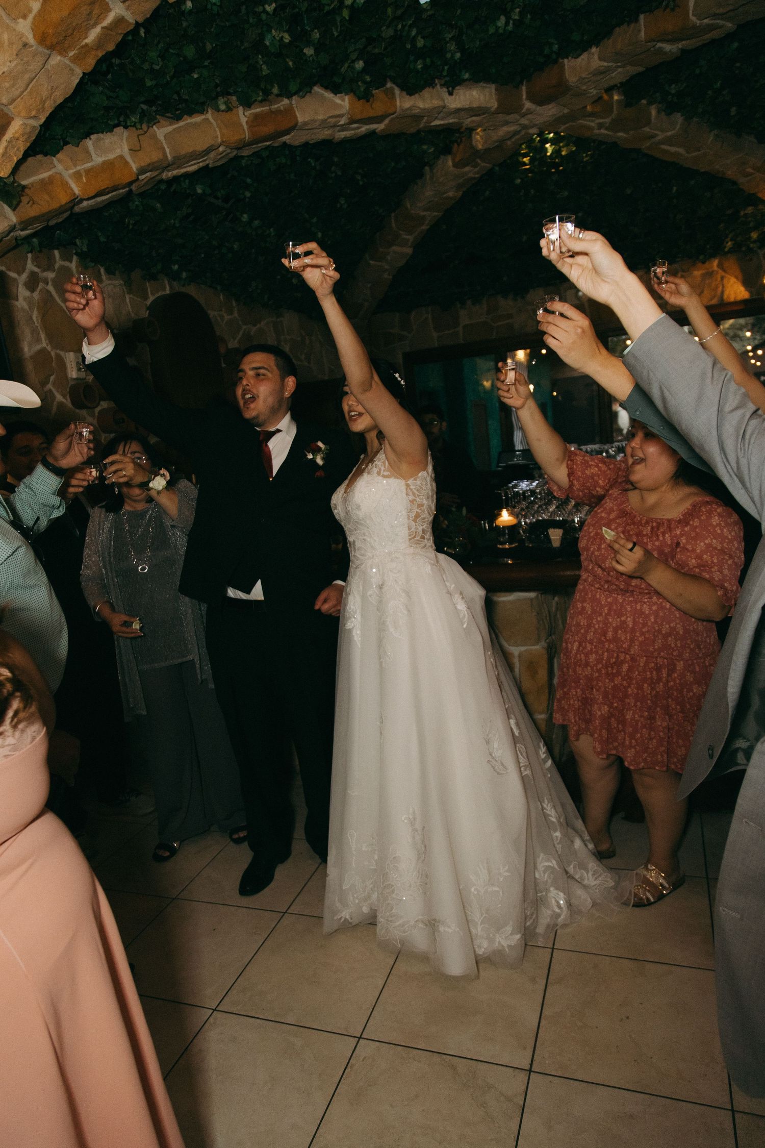 Bride raises her arm triumphantly as guests toast with shot glasses under stone arches at reception.