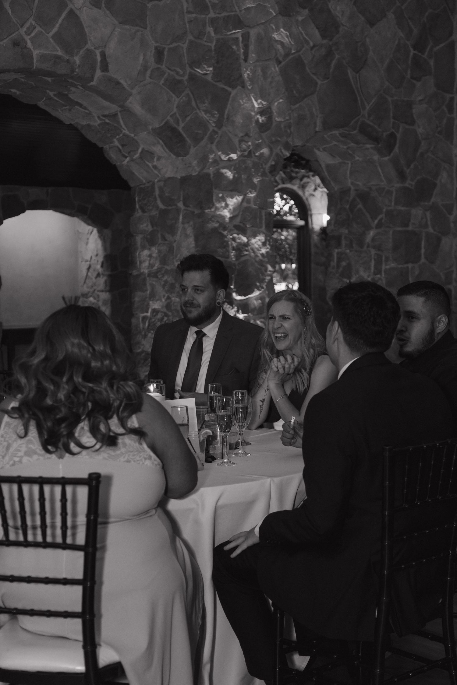 Wedding guests laugh and converse at a candlelit reception table in a stone-walled venue.