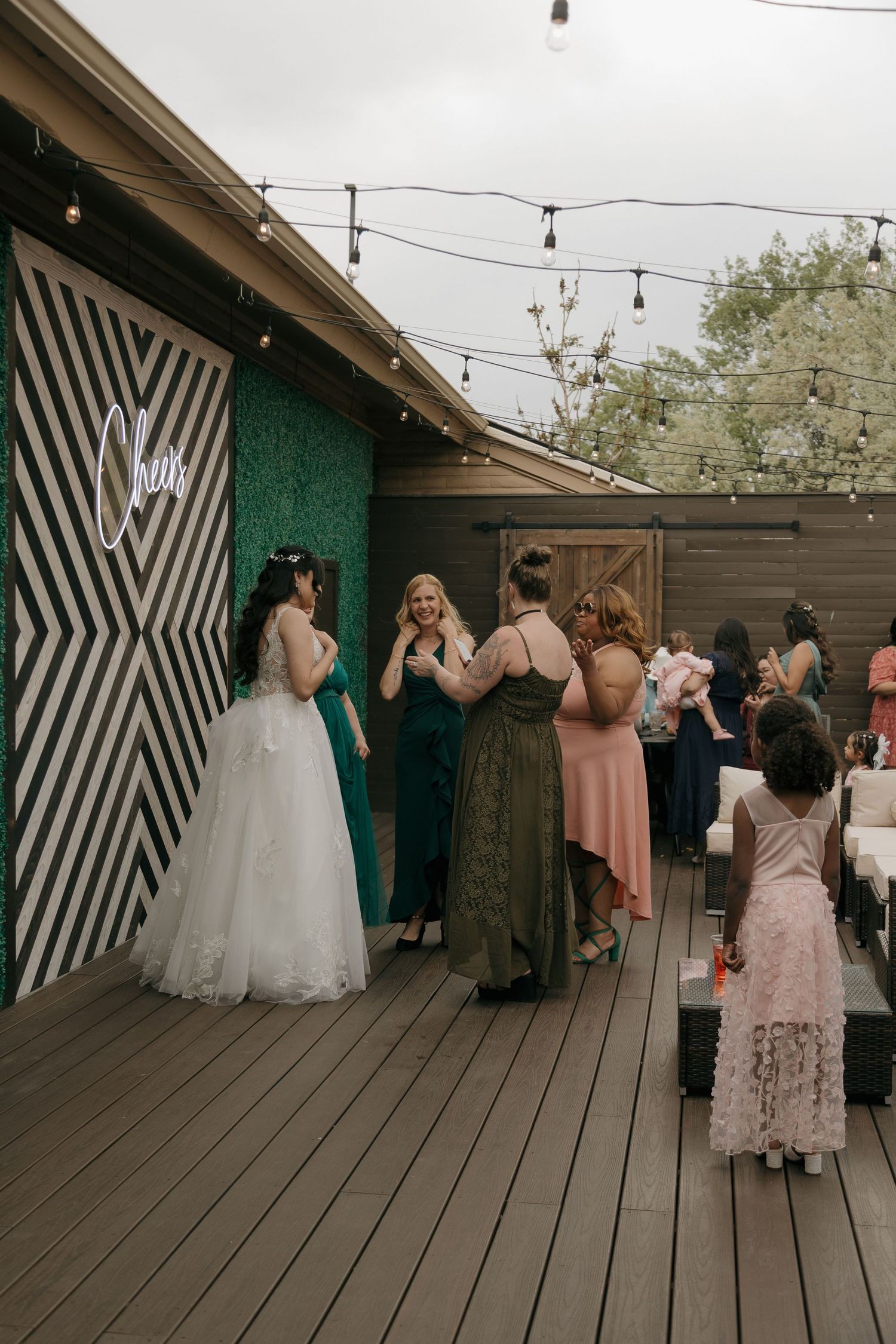 Bride in white gown greets guests near a striped wall and green moss backdrop at outdoor reception.