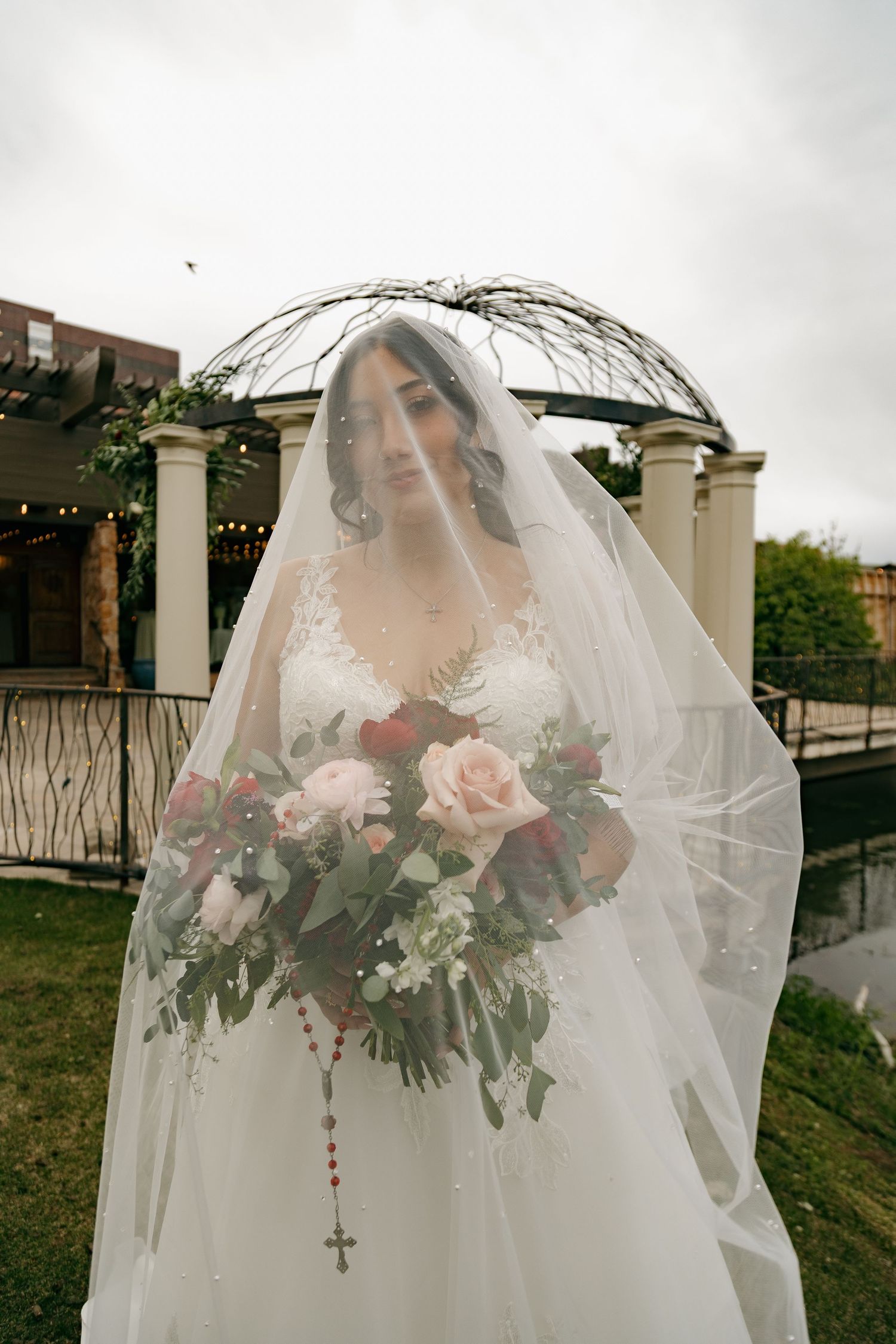 Bride draped in flowing veil holding a lush red and pink rose bouquet near a white outdoor gazebo.