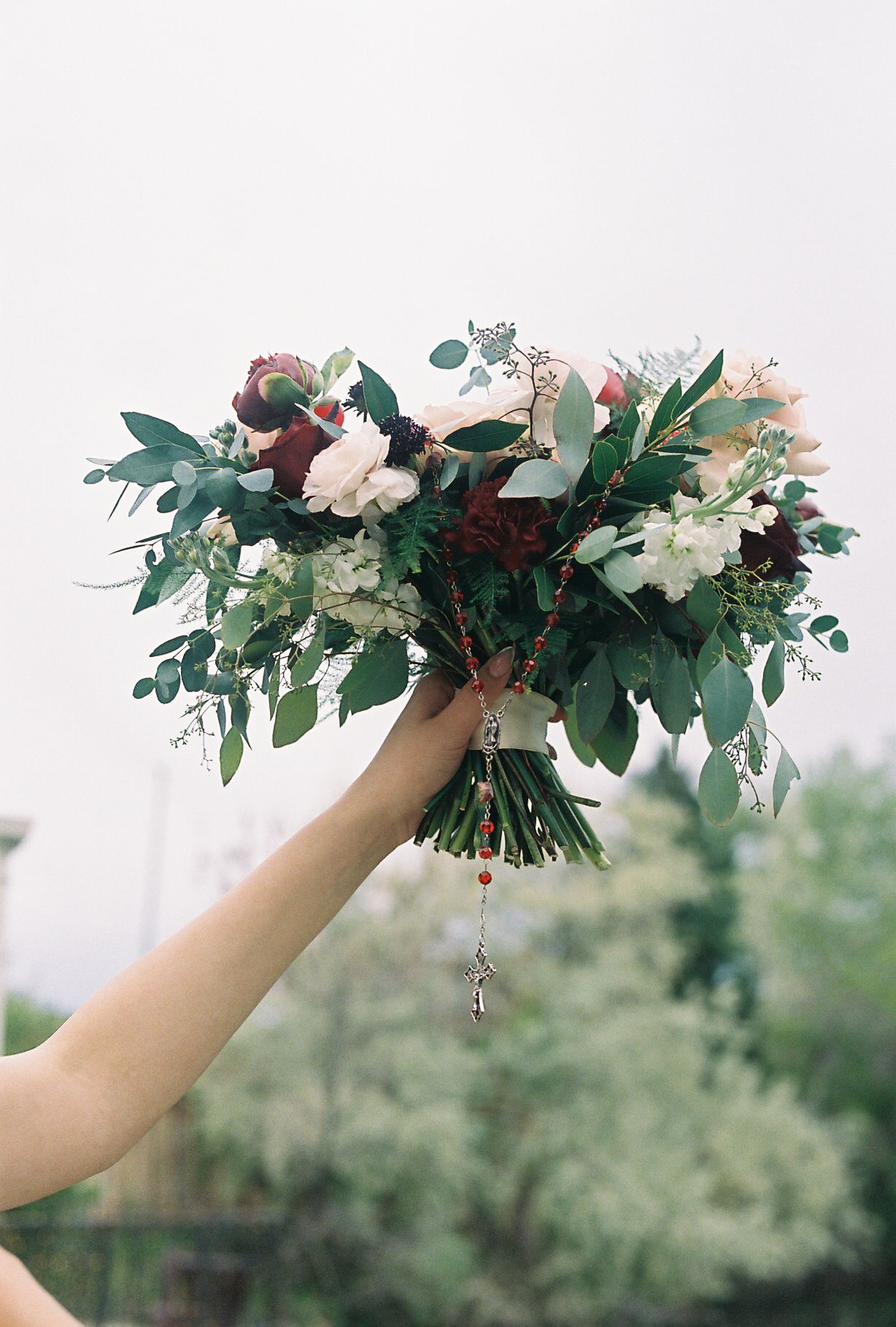 Bride holding lush wedding bouquet of white, red, and green flowers with rosary beads dangling below.