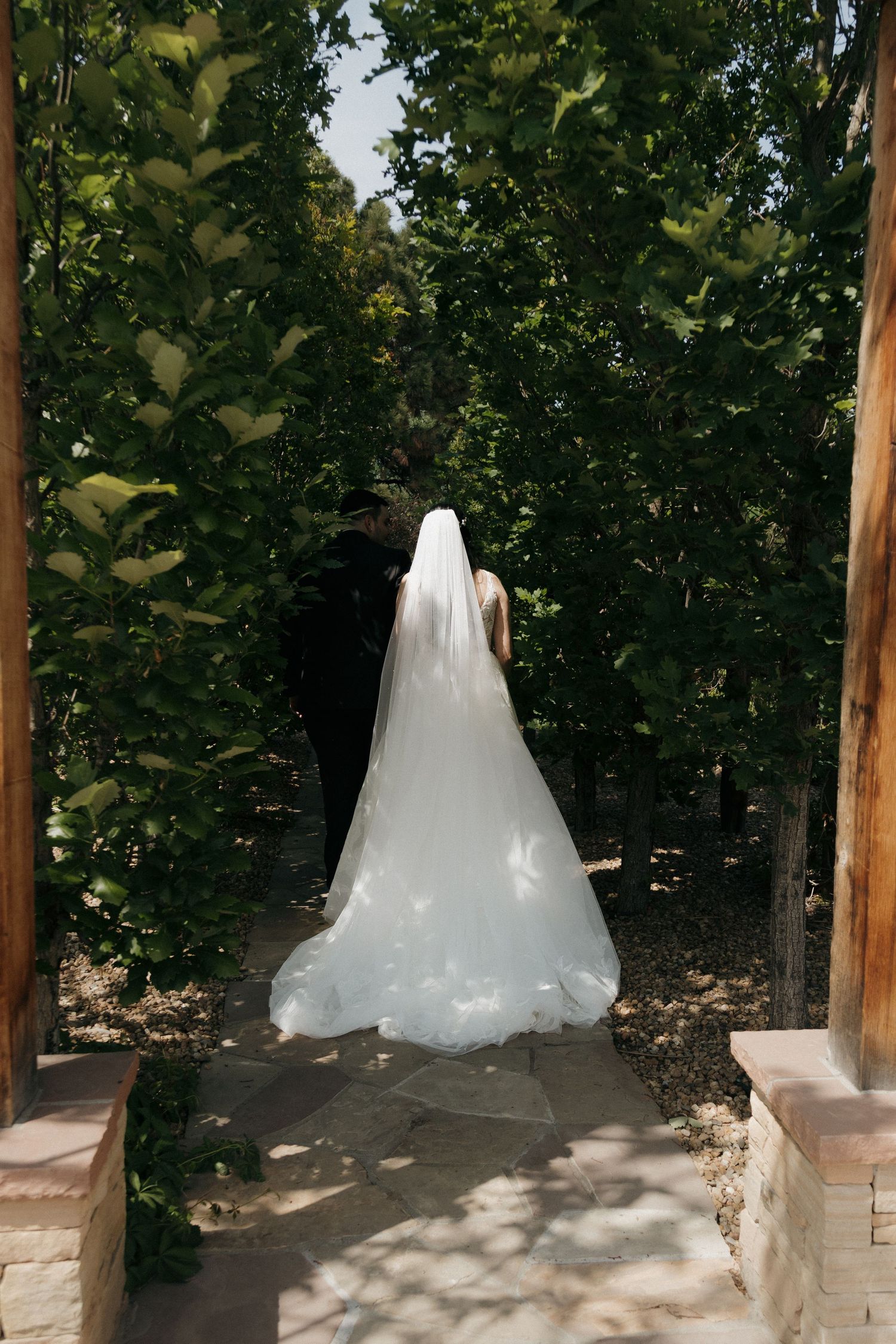 Bride in a long veil and groom walking hand in hand through a sunlit garden archway.