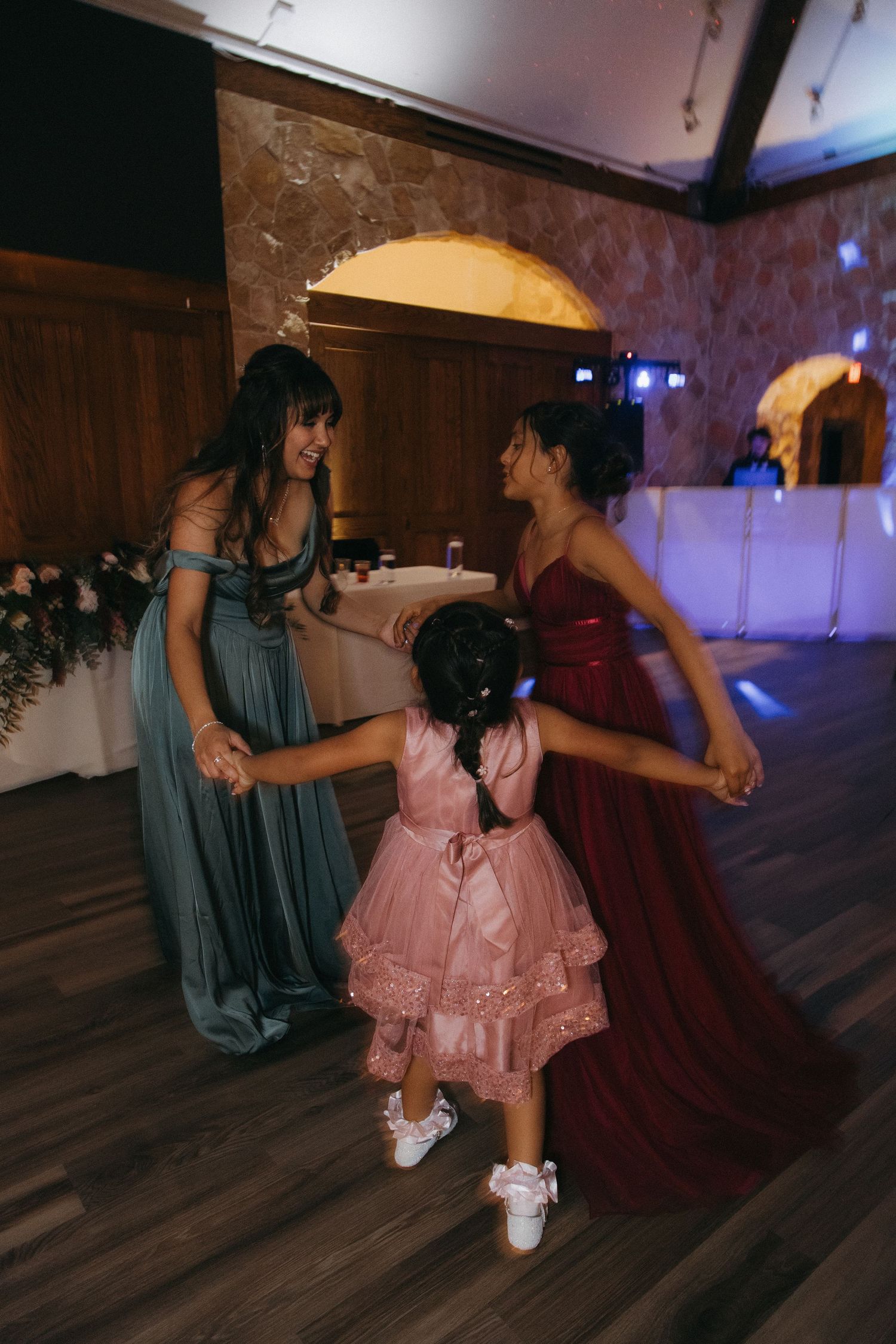 Two bridesmaids in teal and burgundy gowns dancing joyfully with a young flower girl in pink at a wedding reception.