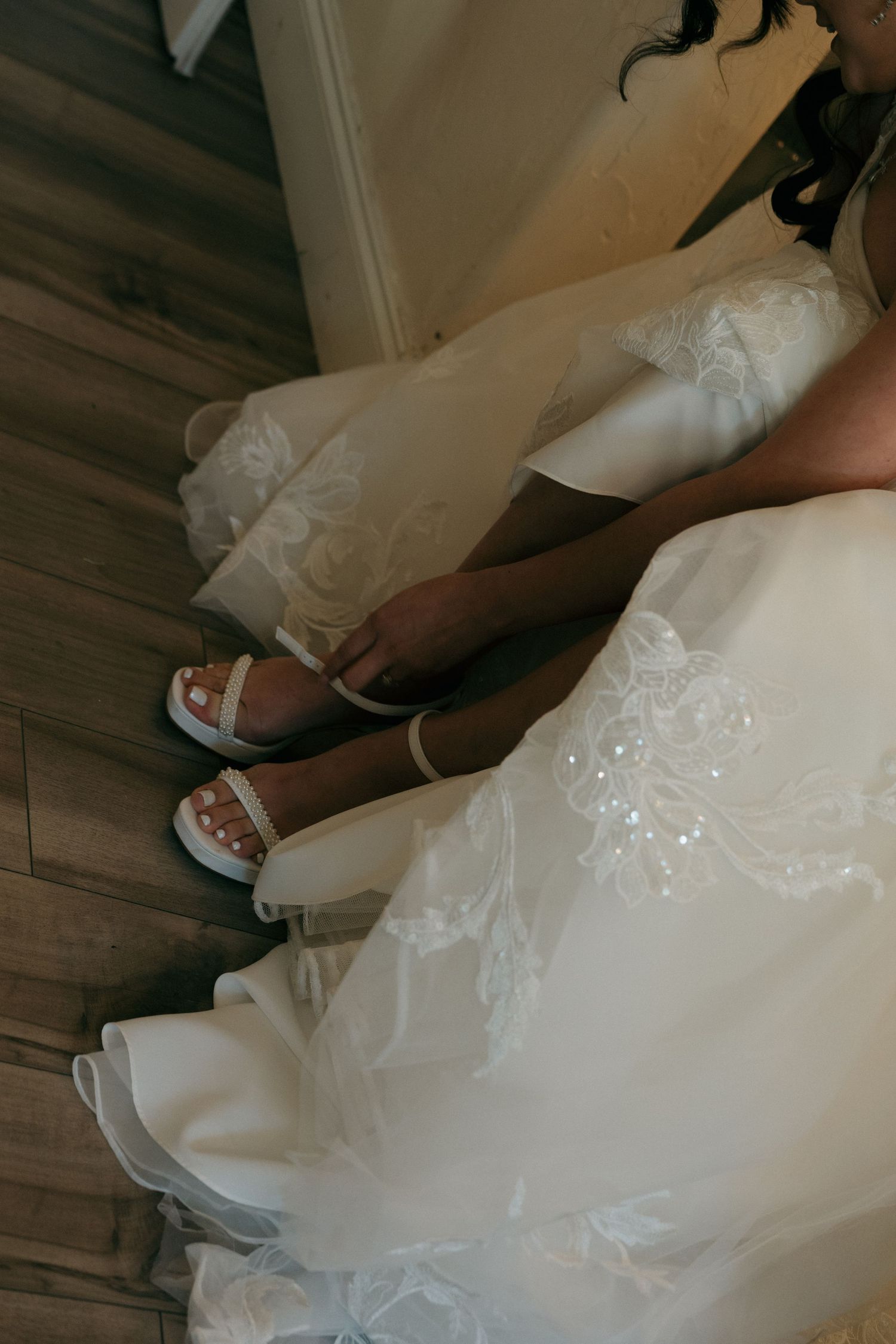 Close-up of a bride in a white lace gown buckling white strappy wedding heels on hardwood floor.