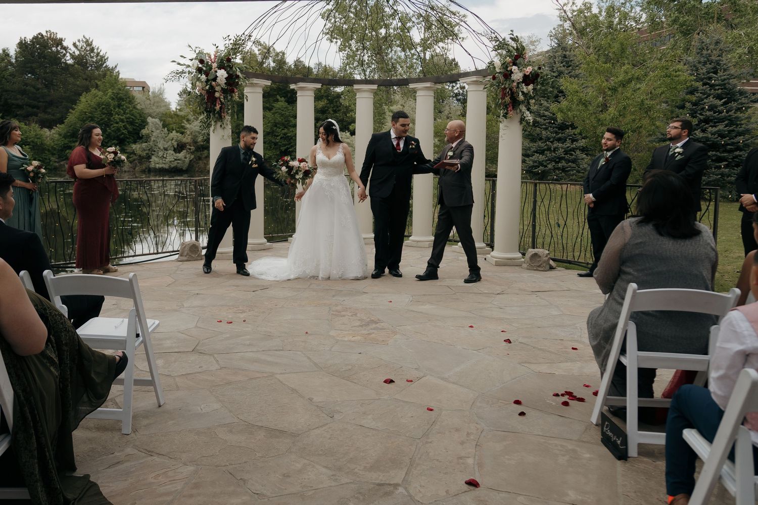 Newlyweds walk hand in hand down aisle after outdoor wedding ceremony by scenic pond with guests.