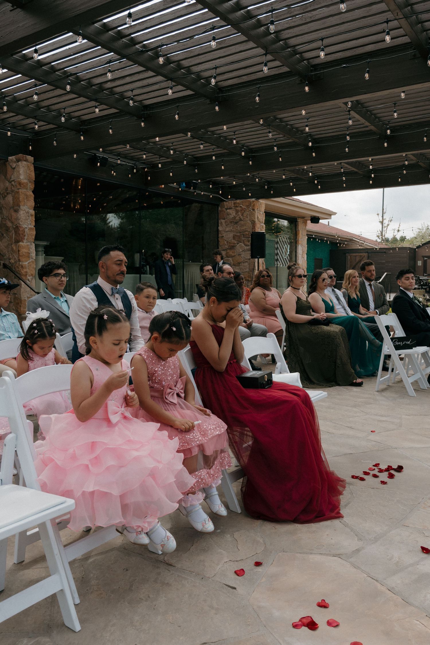 Wedding guests seated outdoors during ceremony, flower girls in pink dresses bow heads in prayer on white chairs.