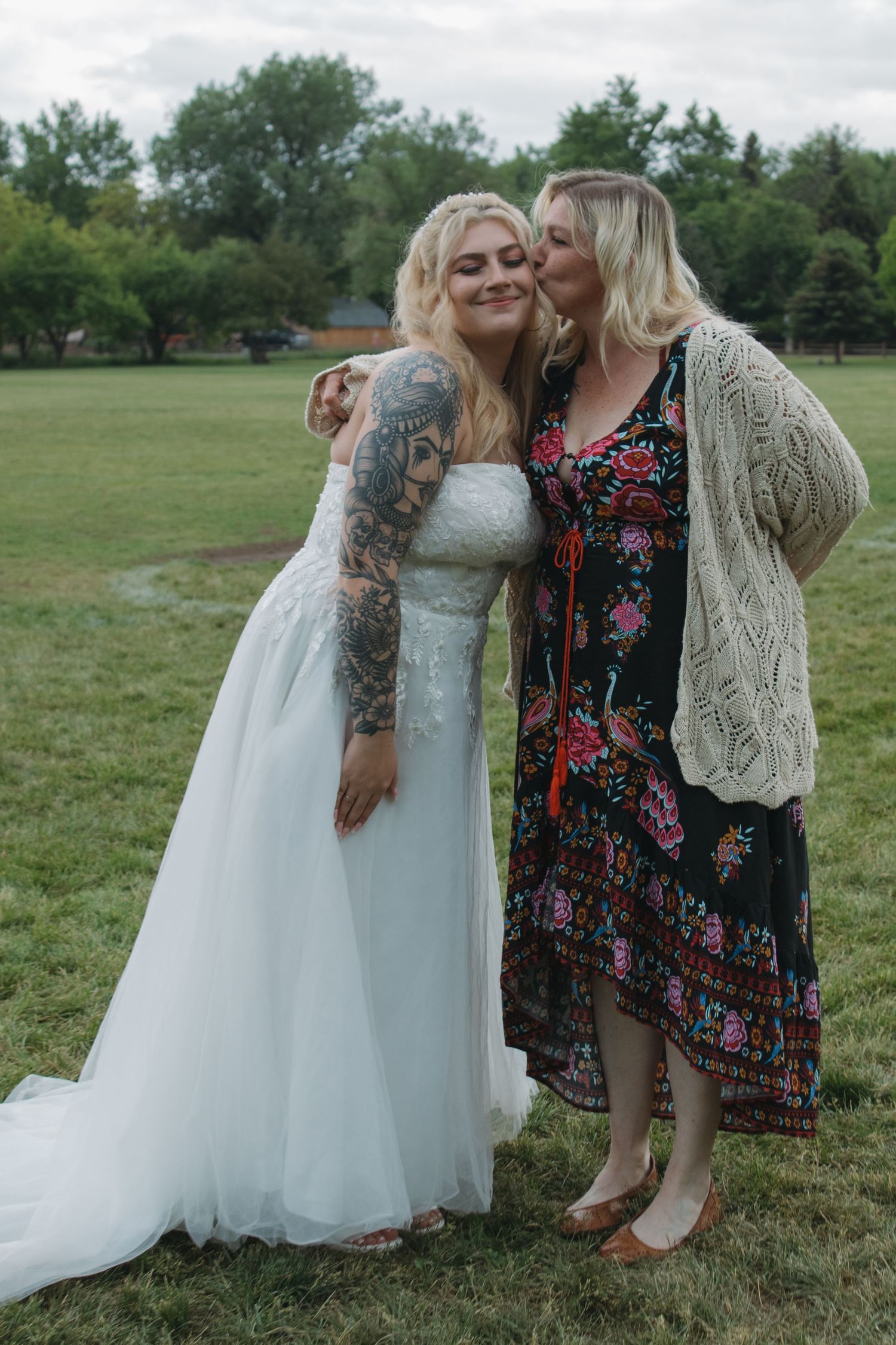 A bride in a white gown with sleeve tattoos stands with a guest wearing a floral dress in an outdoor setting.