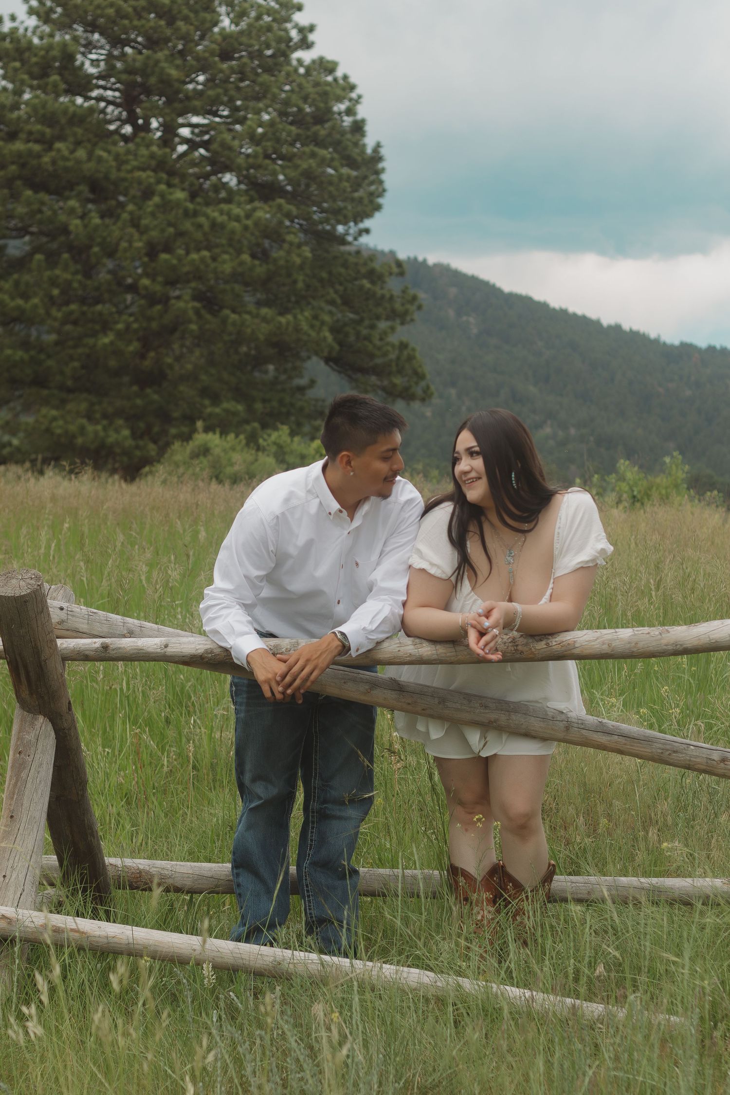 Couple shares sweet moment while resting on rustic fence in mountain grassland setting.