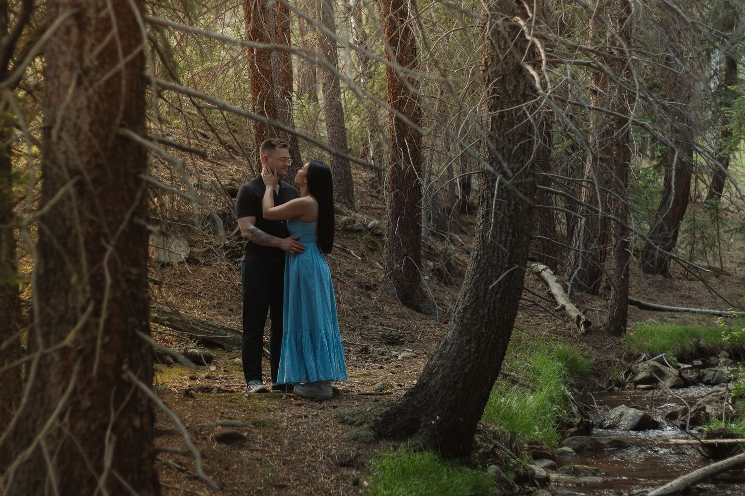 Two people embrace on a winding forest trail surrounded by tall pine trees.