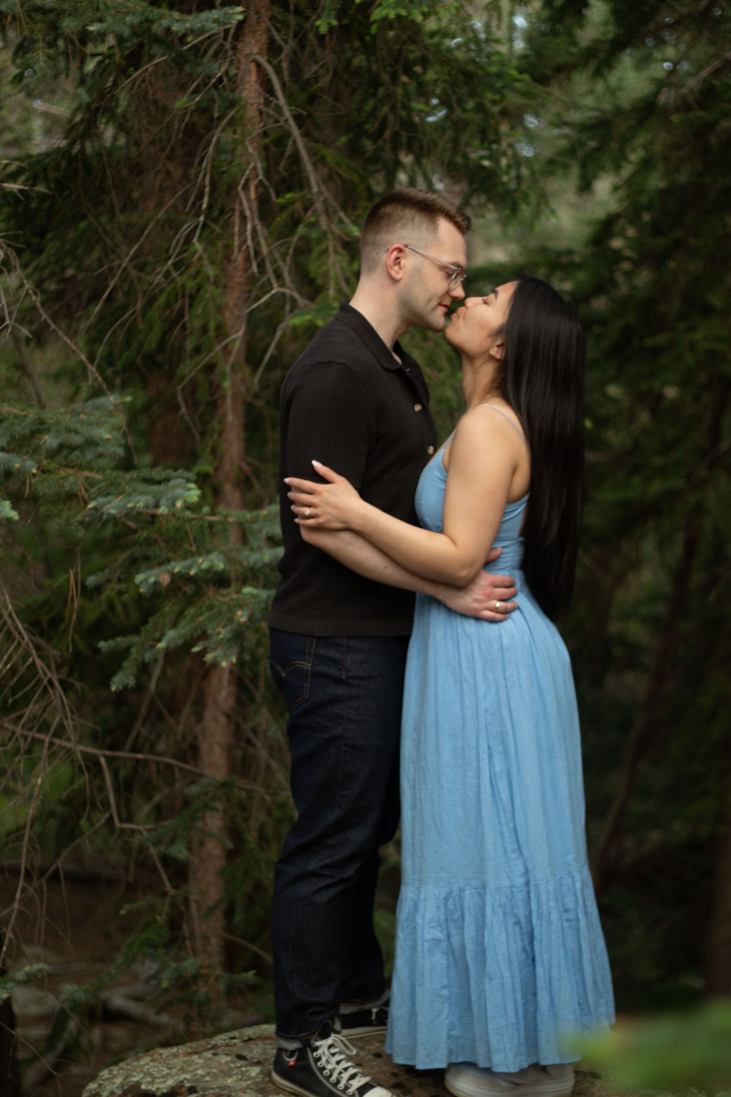 A couple embracing on a rock surrounded by evergreen trees in natural light.