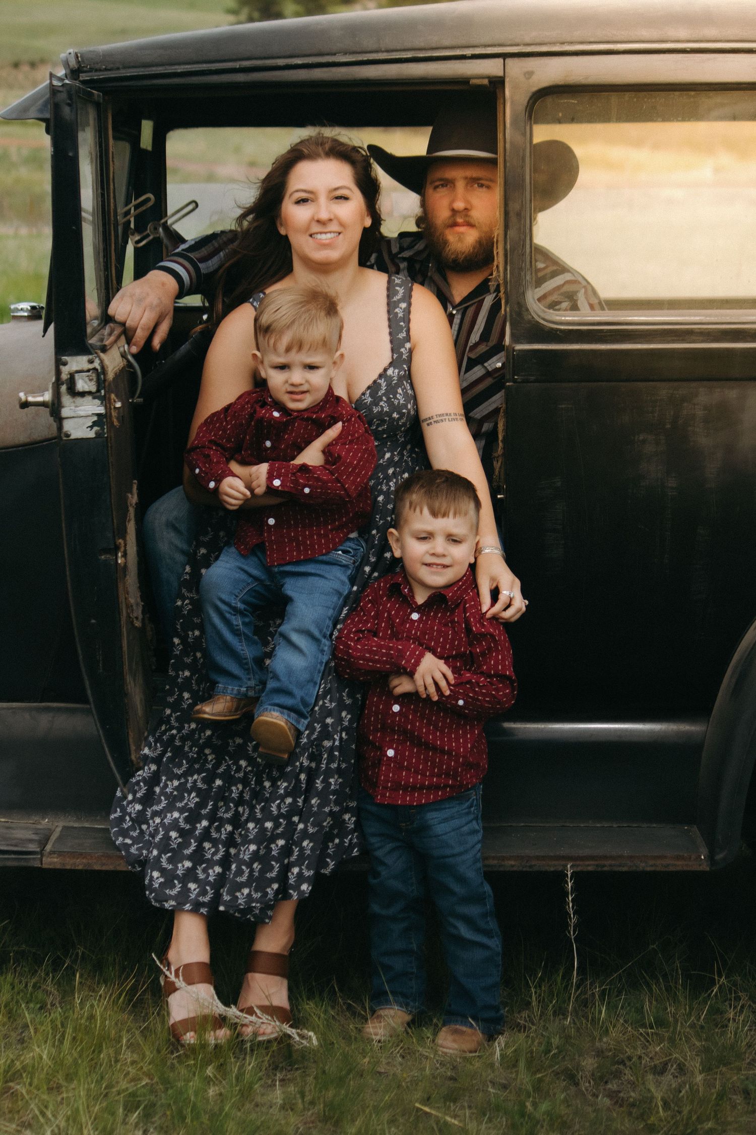 Family posing together at the open door of a vintage black automobile in a grassy field.