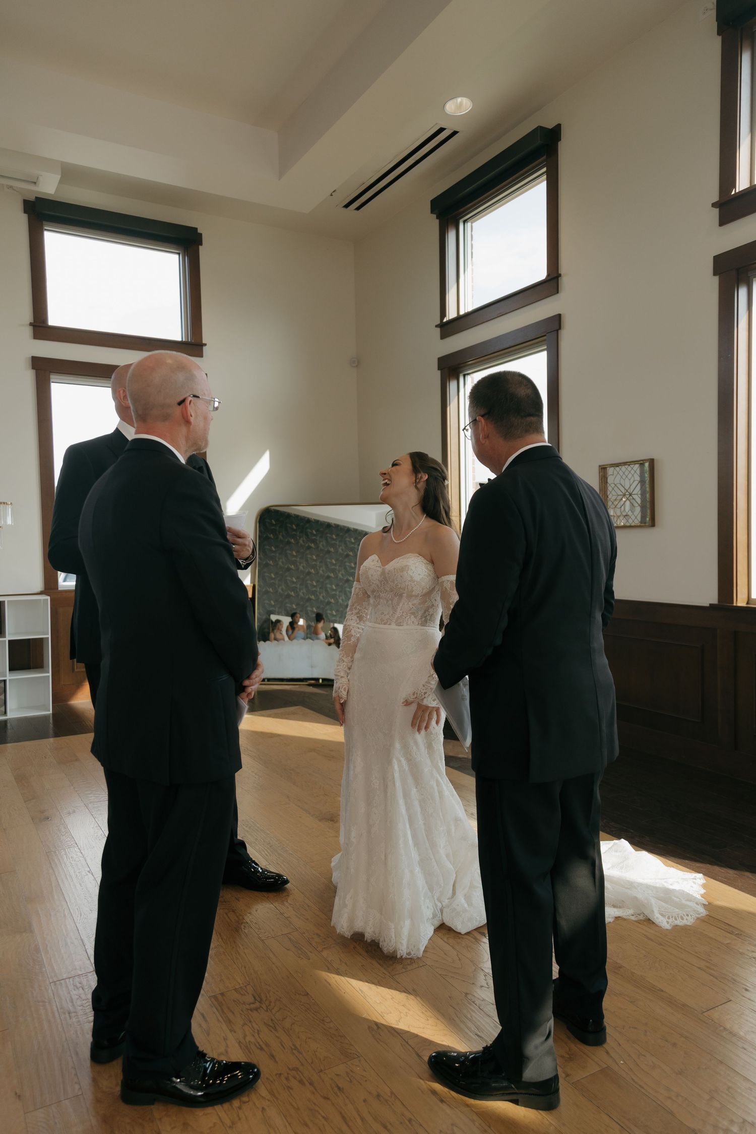 Bride in lace gown smiling during an intimate indoor wedding ceremony with two suited guests.