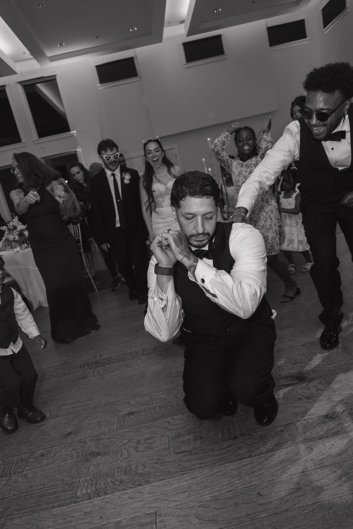Black and white photo of a groom dancing on one knee at a lively wedding reception with guests.