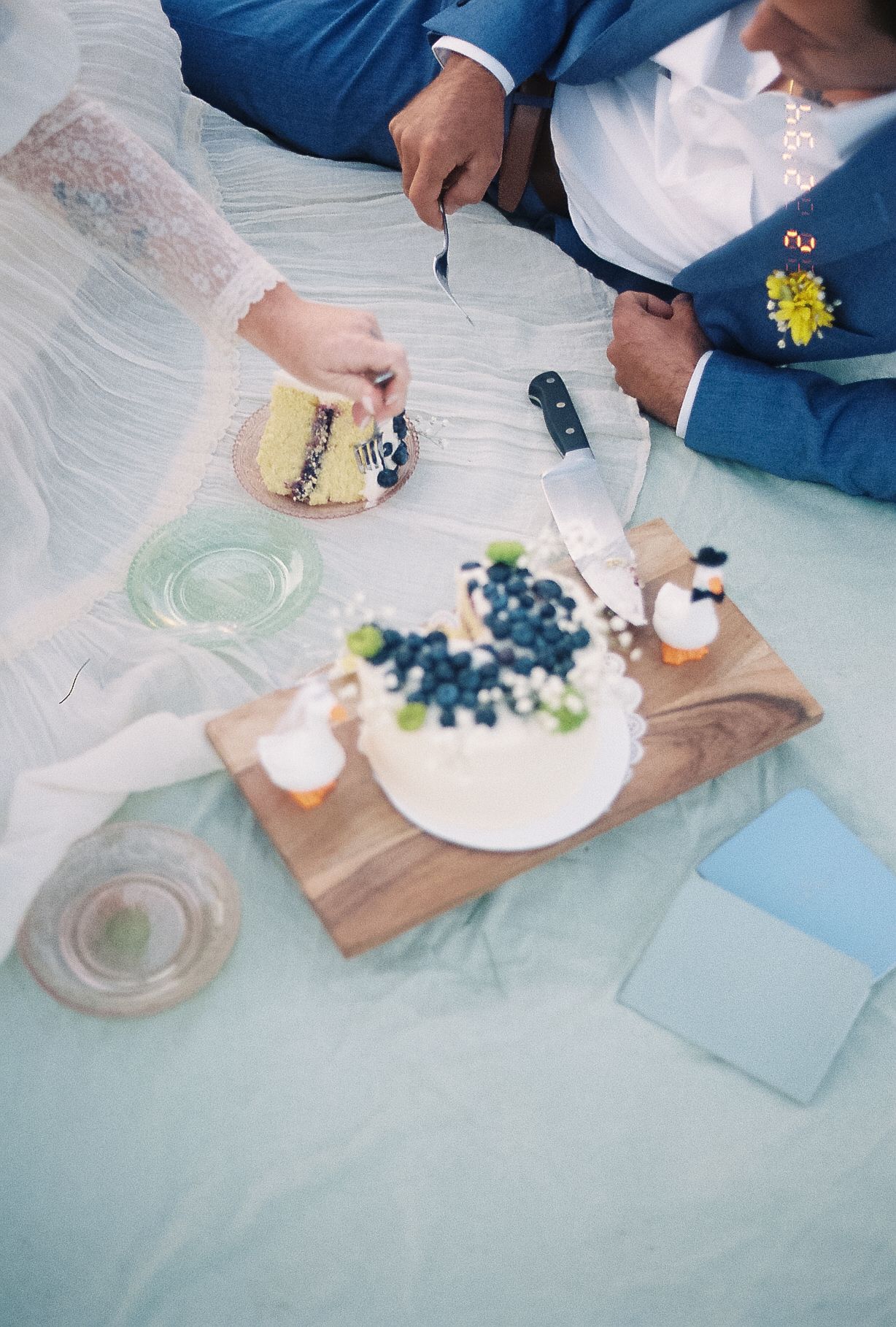 Bride and groom cutting a white wedding cake with blueberries on a wooden board during an outdoor picnic.