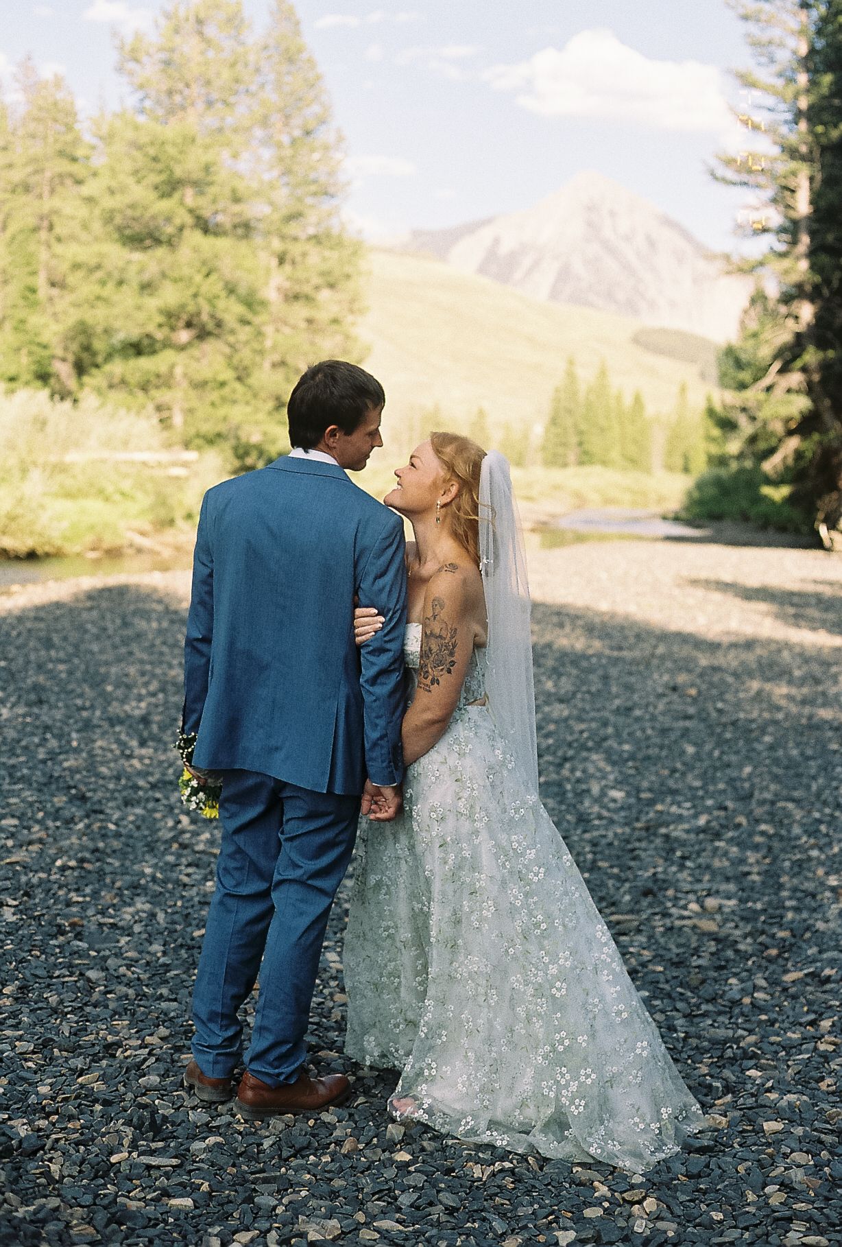 Wedding couple sharing intimate moment on rocky path, bride in floral lace gown with mountain views.