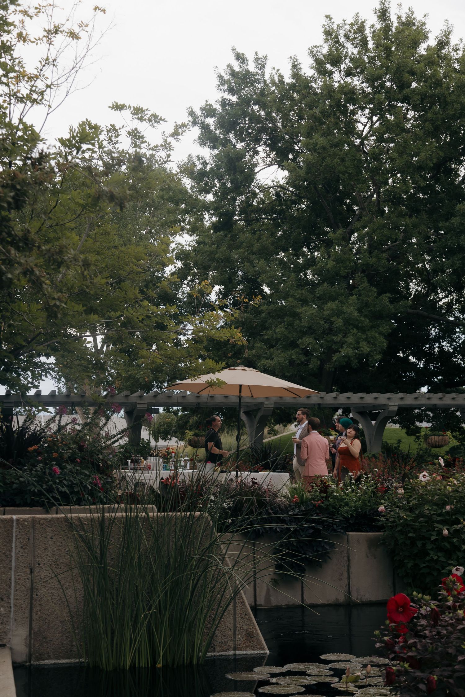 Outdoor garden wedding venue with guests mingling near pergola, flowers, and pond under overcast sky.
