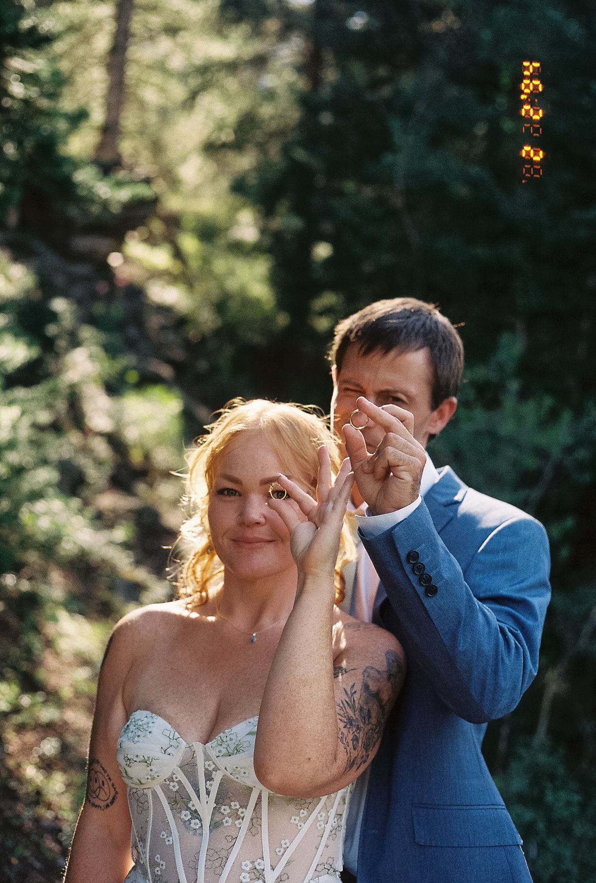Bride and groom making heart shape with fingers in sunlit forest, bride wearing lace corset wedding dress.