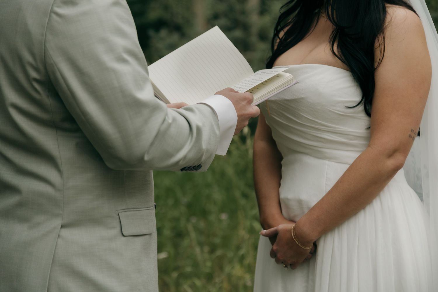 Groom in grey suit reads wedding vows while holding bride's hands outdoors.