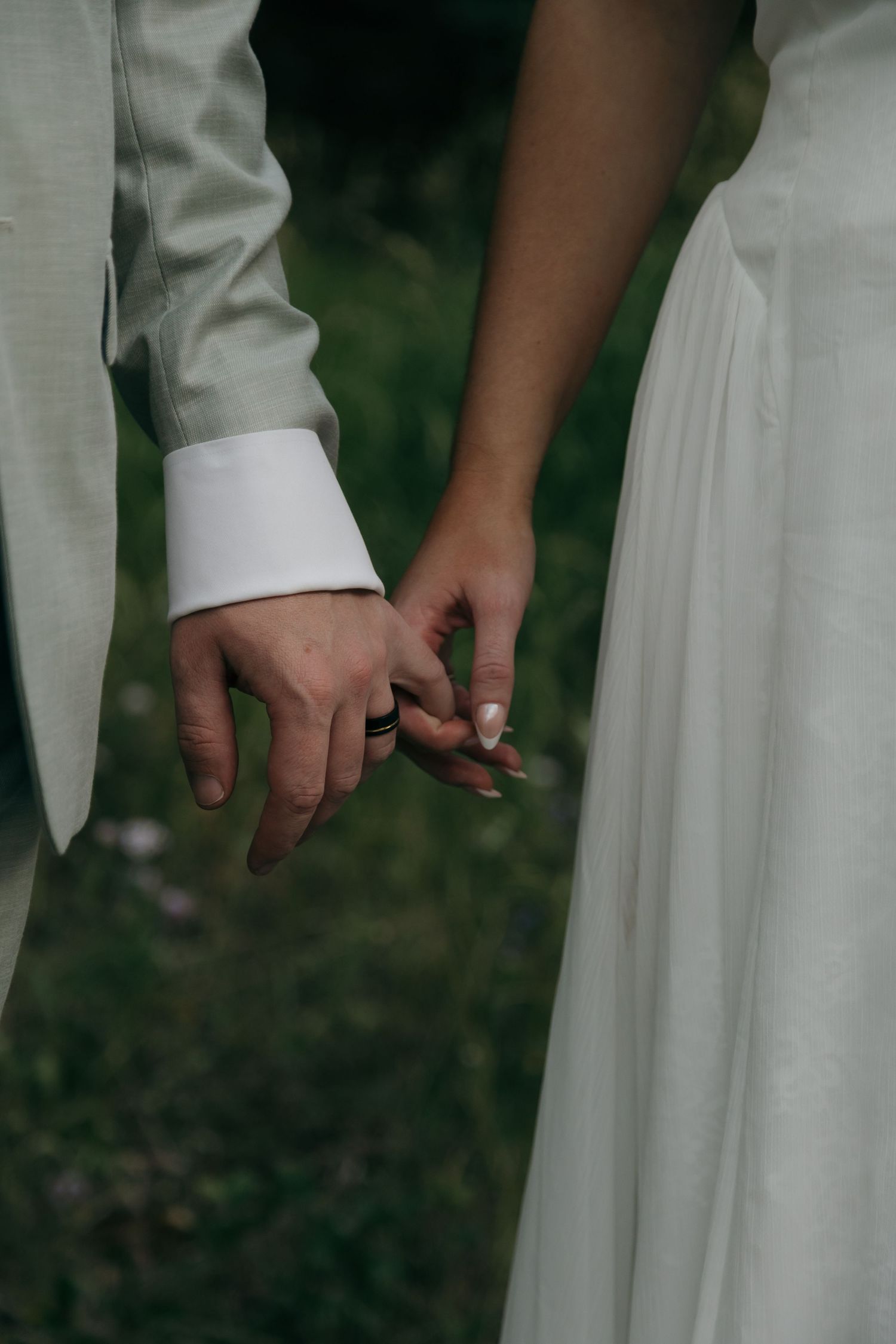 Close-up of bride and groom gently holding hands outdoors, groom wearing a wedding ring.