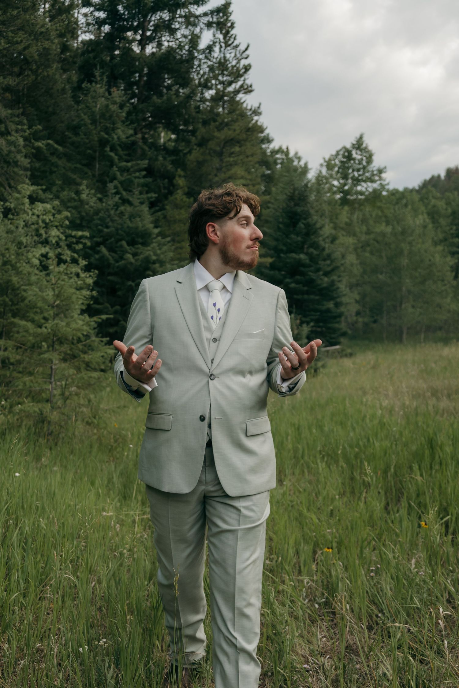 Groom in sage three-piece suit stands confidently in a lush green forest meadow on wedding day.