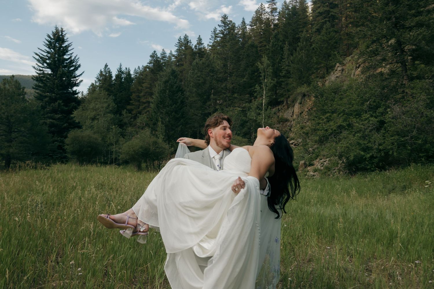 Groom carries laughing bride in white gown through green mountain meadow on their wedding day.