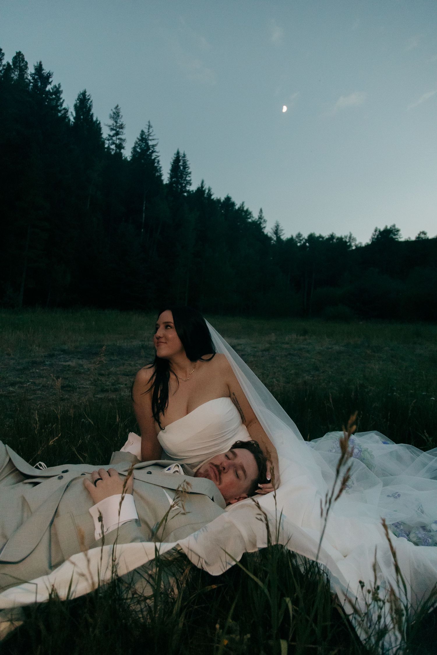 Bride in white gown sitting with groom lying in her lap in a grassy field under a crescent moon.
