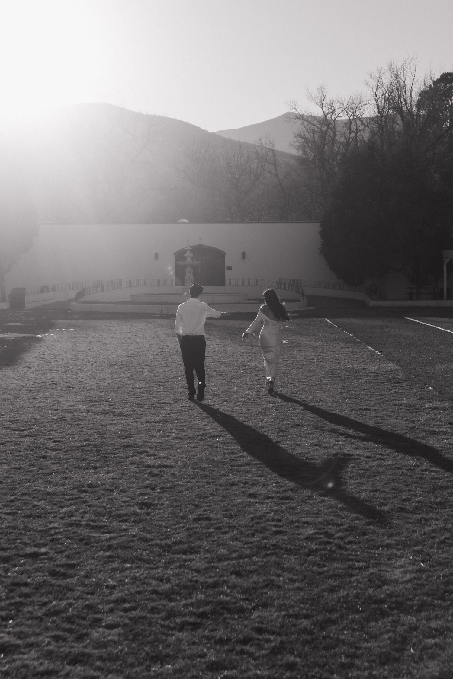 Two silhouettes run together across a field at sunset with mountains in the background.
