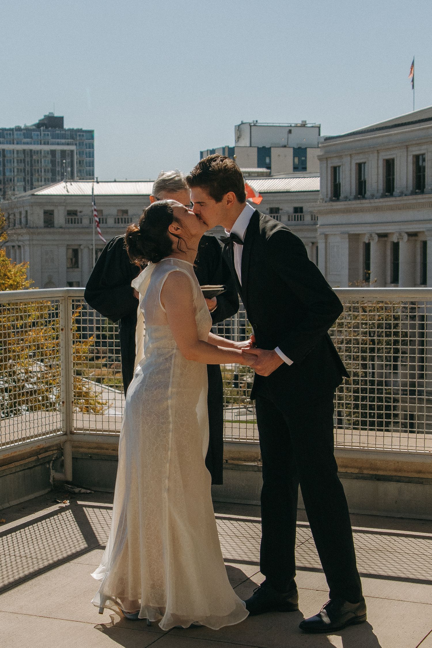 A couple shares a romantic kiss on an outdoor terrace with city buildings in the background.