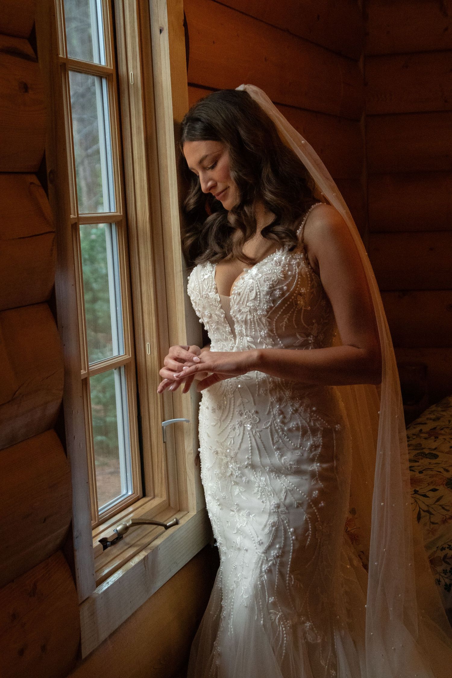 A bride in an elegant lace wedding dress stands by a window in a rustic cabin.