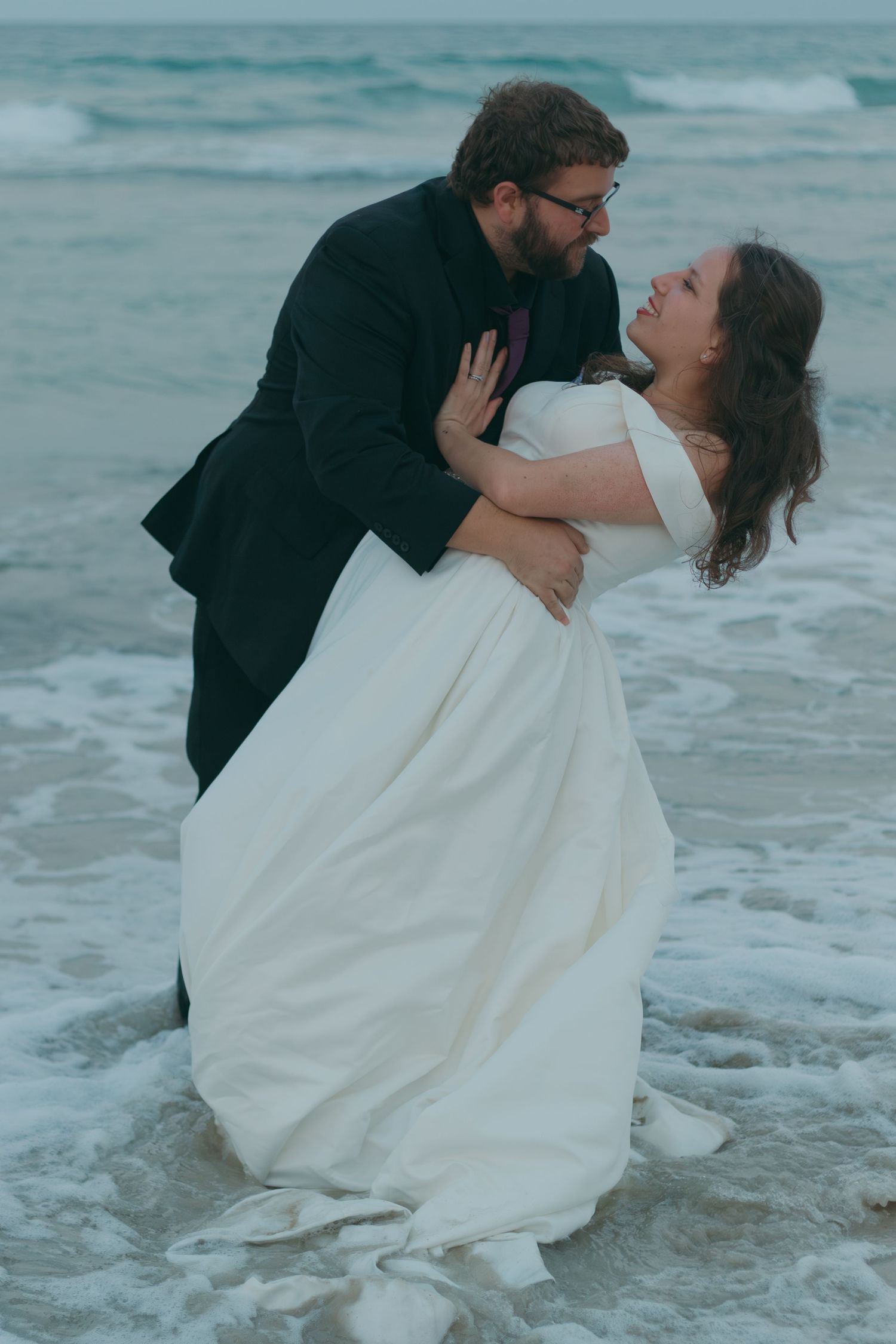 A joyful wedding couple shares a romantic dip pose on the beach with waves and cloudy skies in the background.