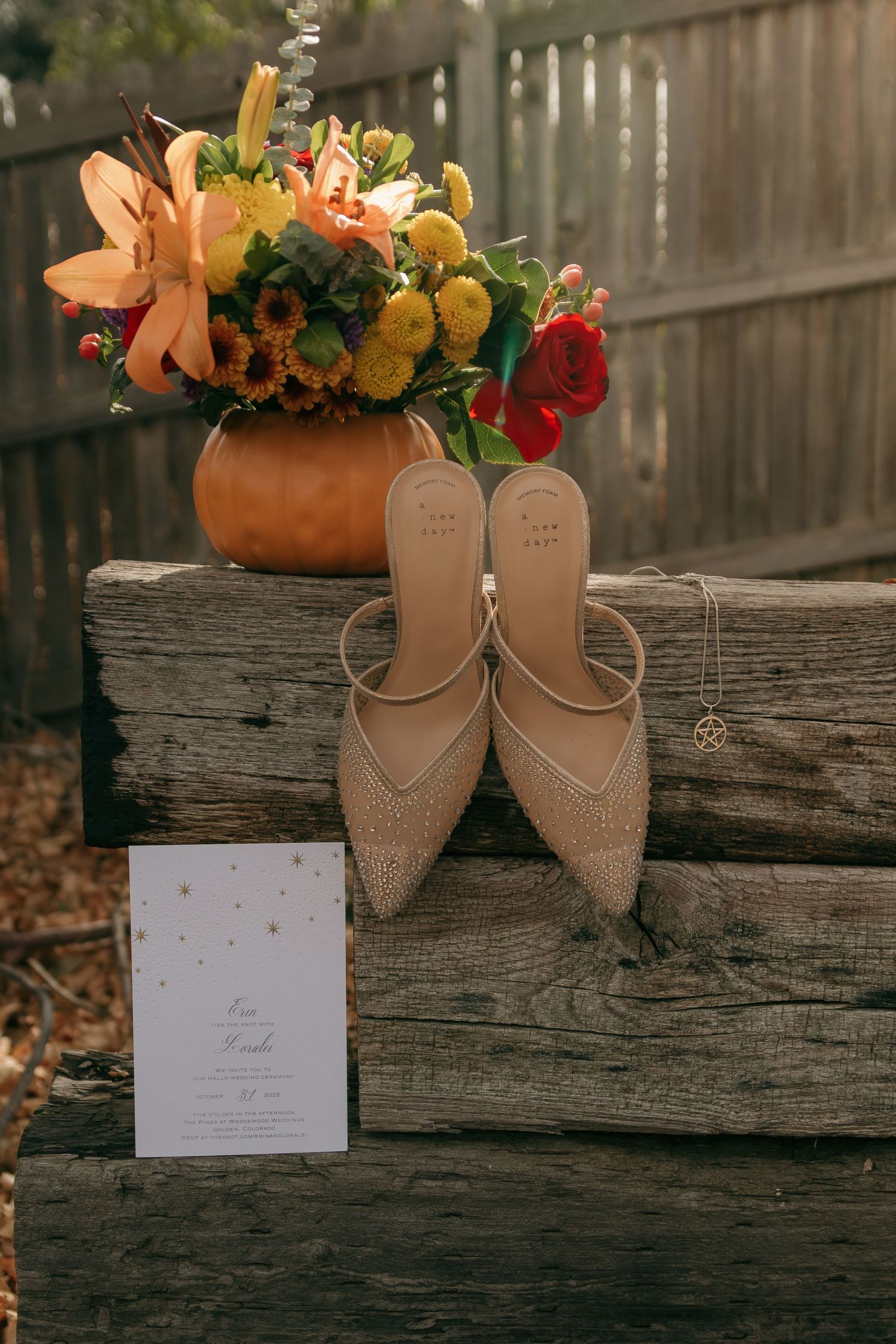 Wedding details featuring beige bridal shoes, autumn floral arrangement in a pumpkin vase, and invitation card on rustic wood.