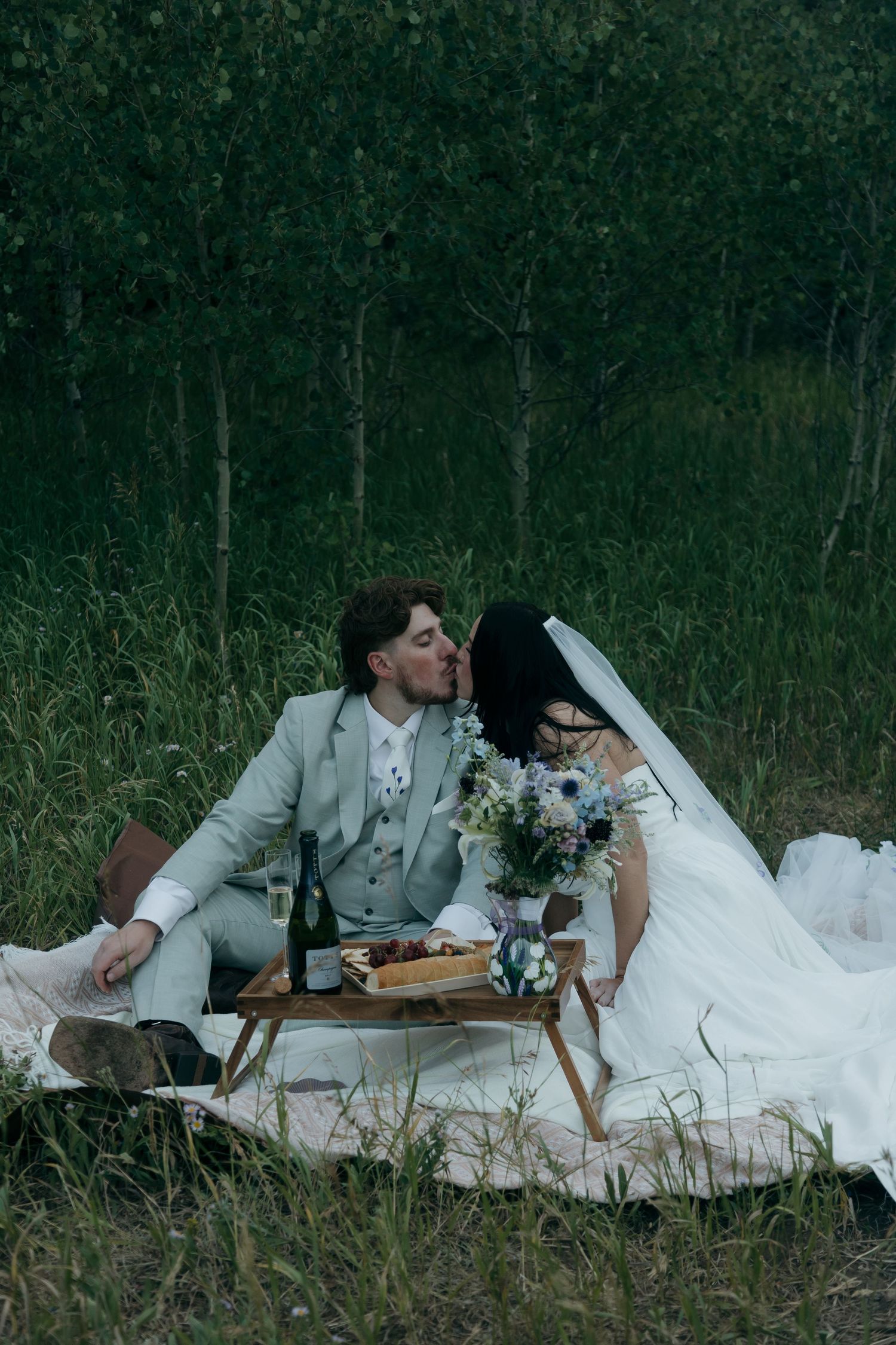 Newlyweds share a kiss during outdoor picnic with wine and flowers in a forest meadow.
