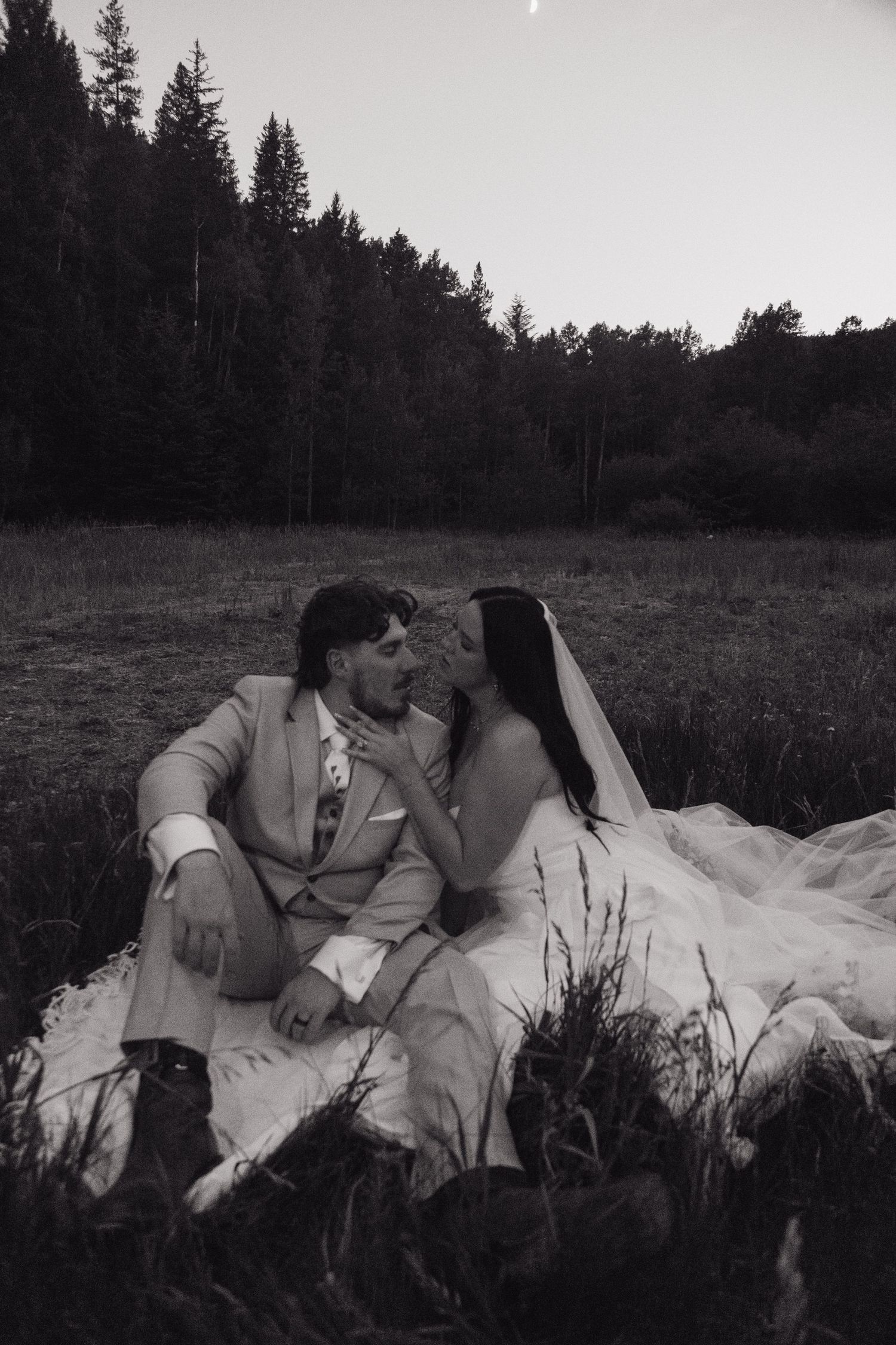Black and white romantic photo of bride and groom sitting together in a meadow at dusk with forest behind.
