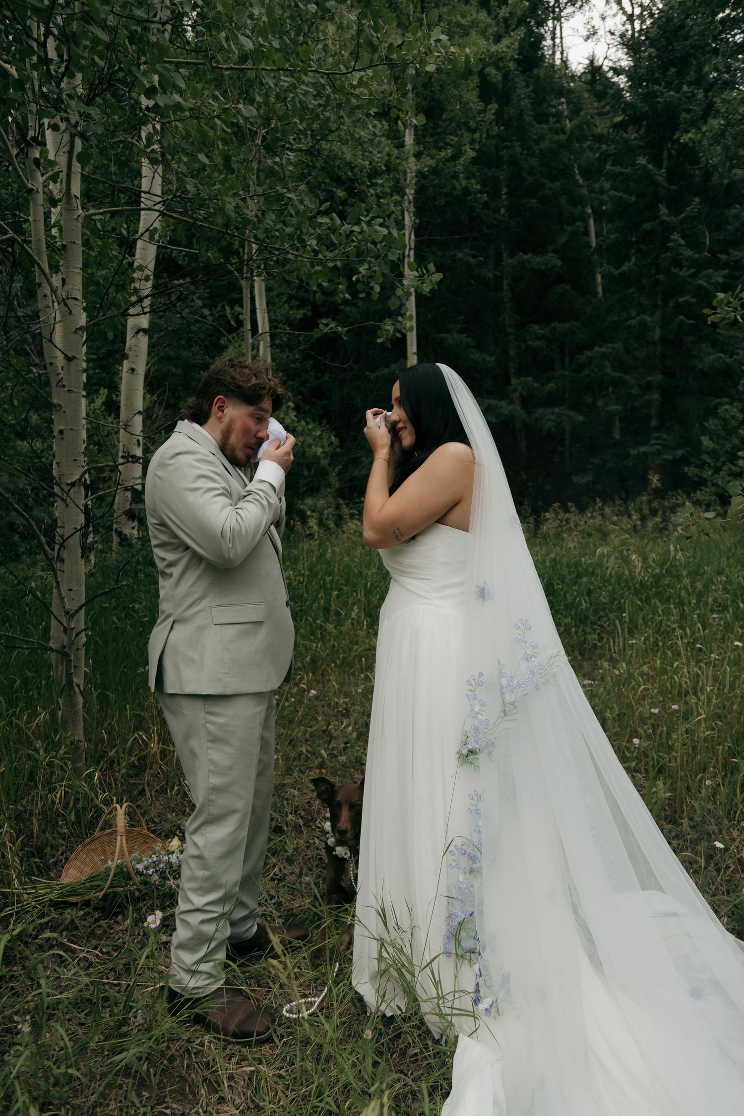 Bride and groom wipe tears during heartfelt outdoor forest elopement ceremony with dog nearby.