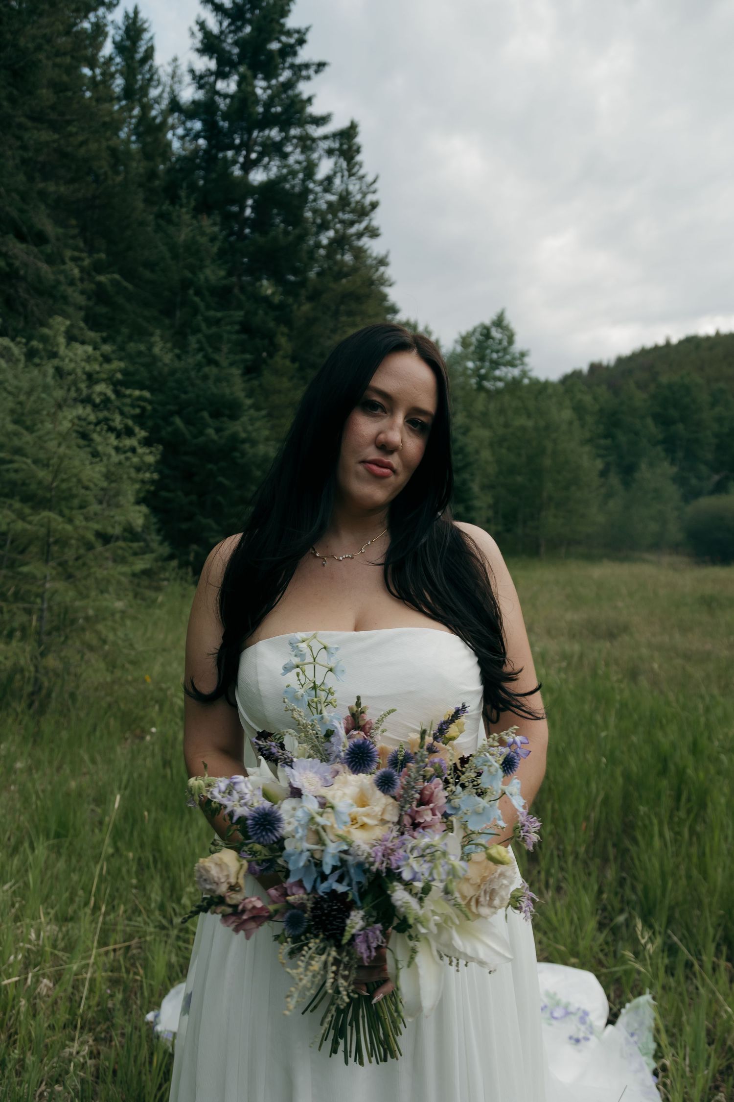 Dark-haired bride in strapless white gown holds a wildflower bouquet in a mountain meadow setting.