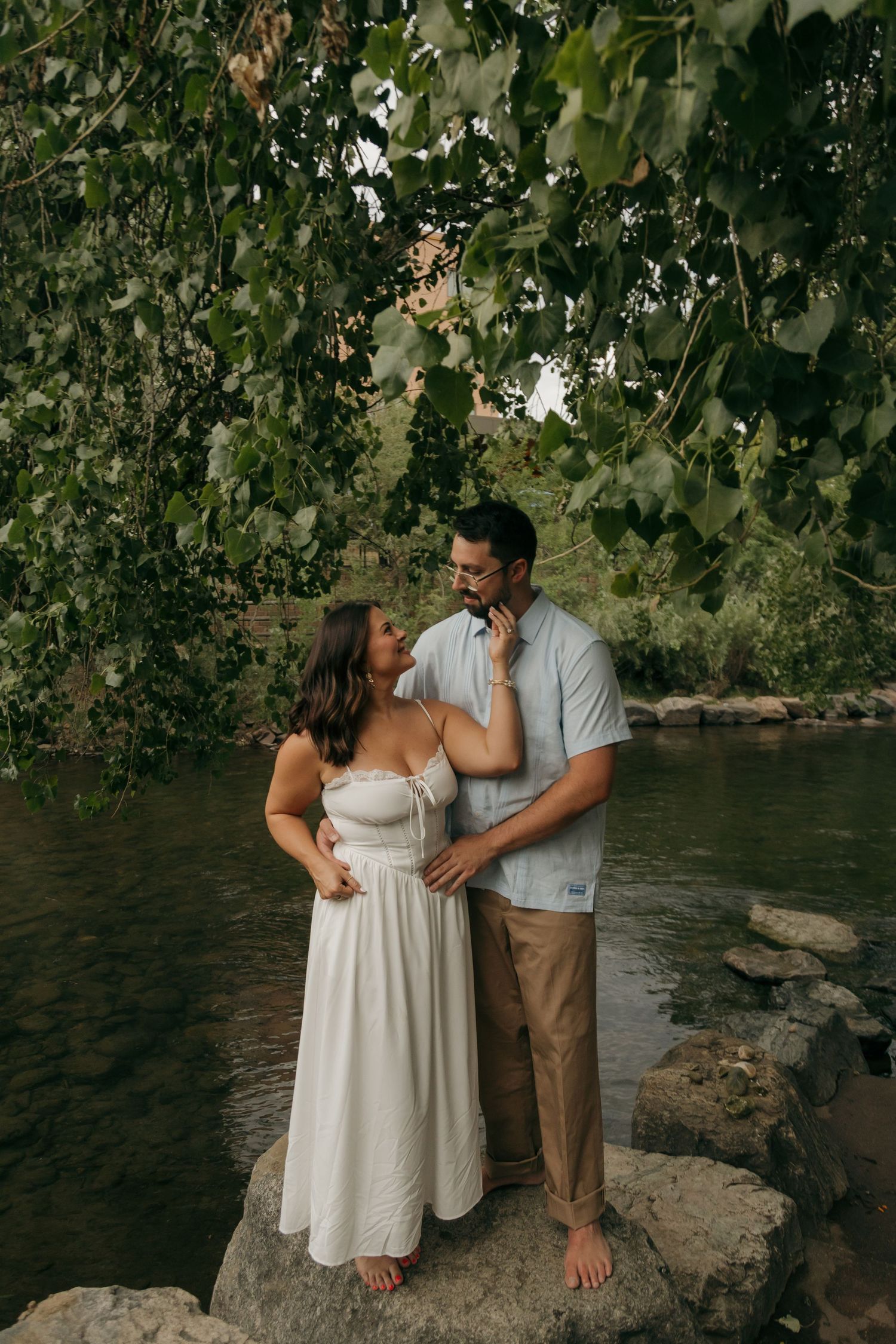 Couple standing on rocks by a river under lush green trees, woman in white dress, man in light blue shirt.