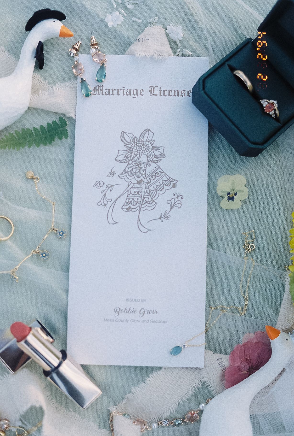 Flat lay of marriage license, wedding rings, swan figurines, jewelry and flowers on sage green fabric.