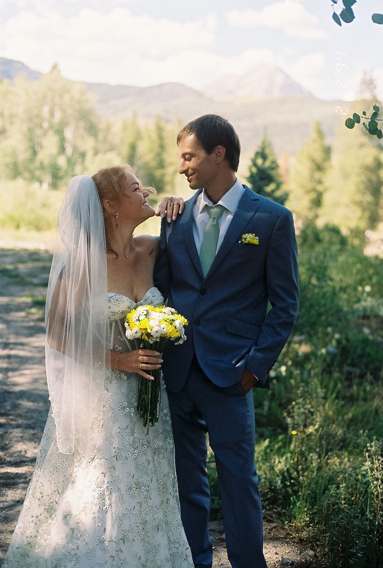 Bride and groom sharing a joyful moment outdoors with mountain views, veil, and yellow wildflower bouquet.