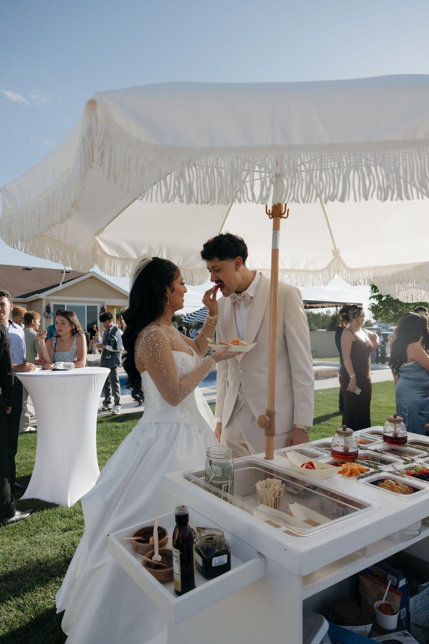 Bride feeds groom a bite of food at outdoor wedding cocktail hour under white fringe umbrella.