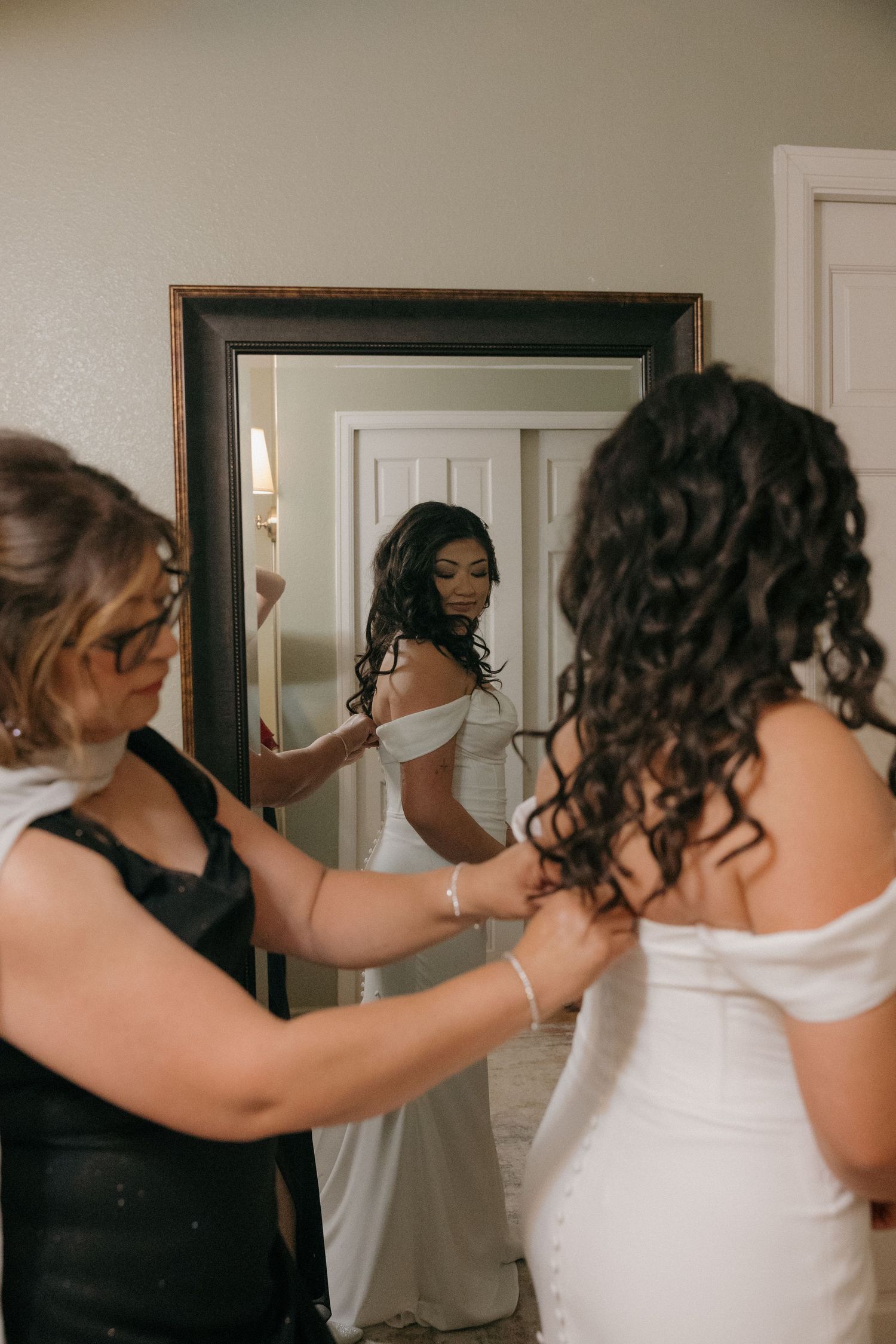 Bride in white off-shoulder gown being helped by two women while standing before a large mirror.