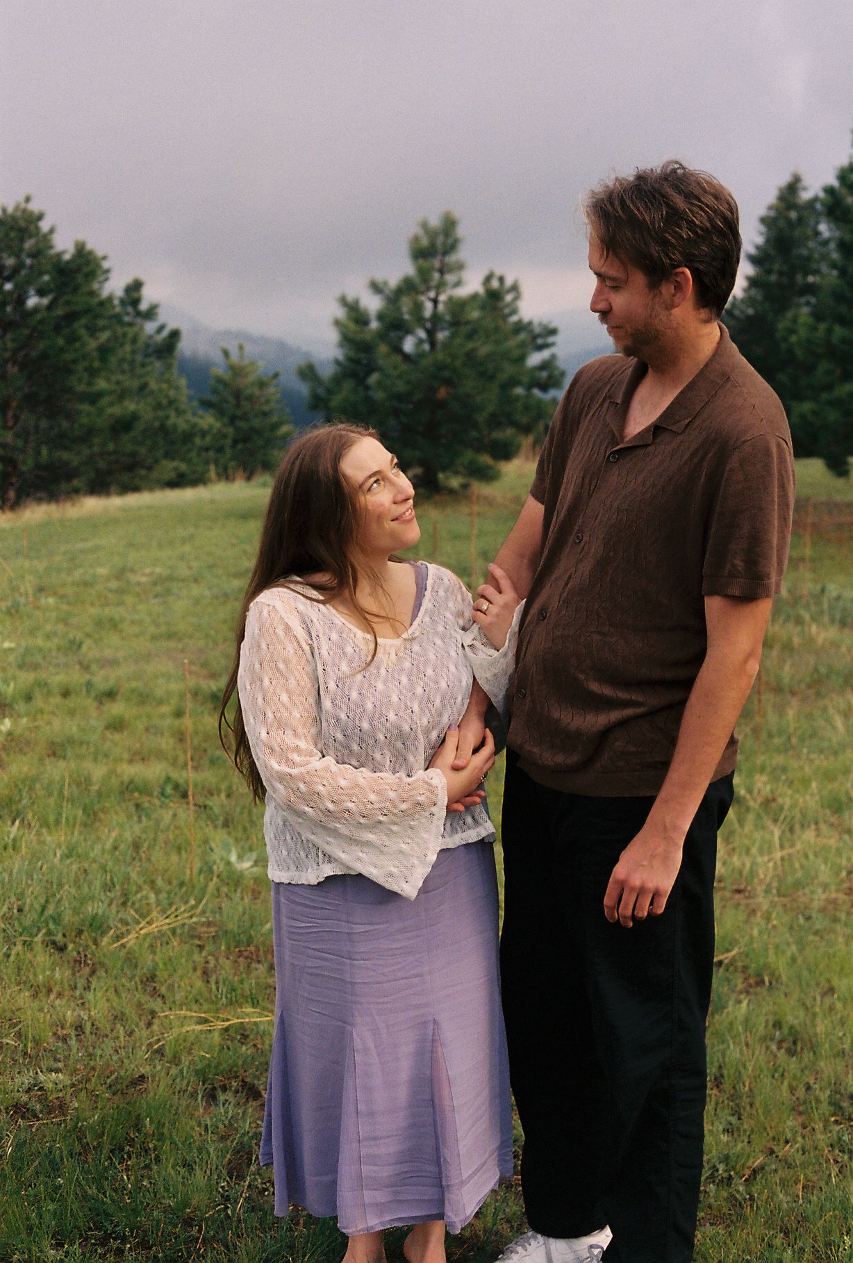 Smiling couple standing together in a green meadow with mountains and pine trees in background.