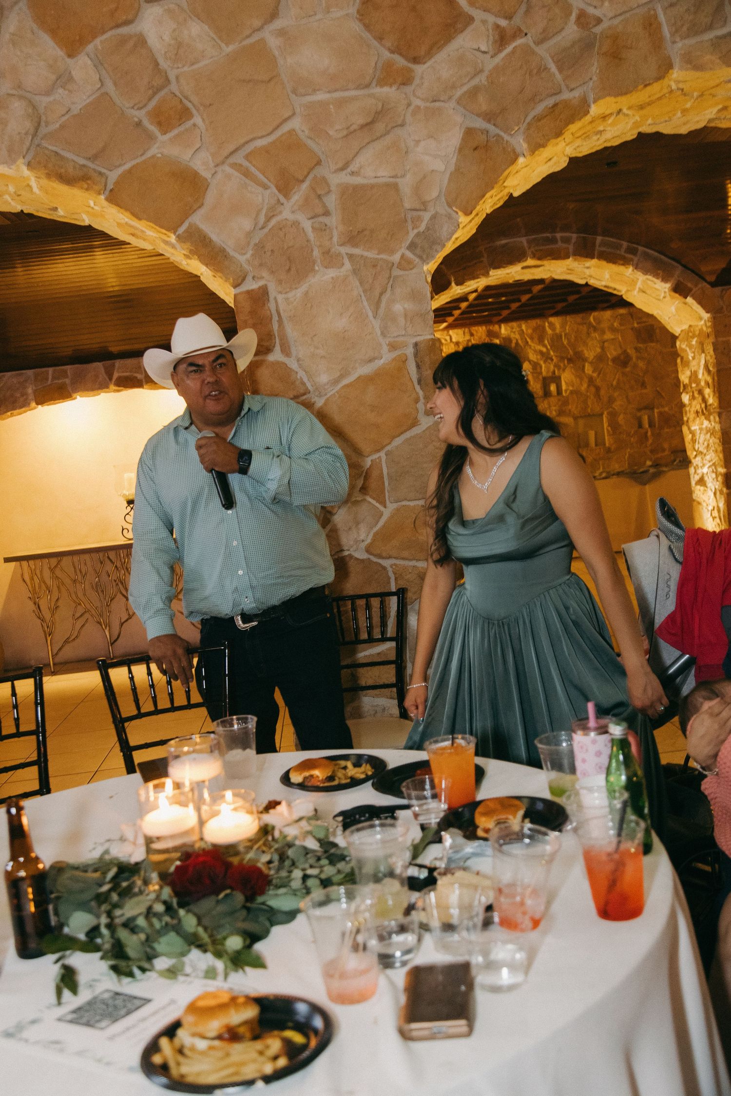 Man in cowboy hat speaks into microphone beside a woman at a candlelit wedding reception table.