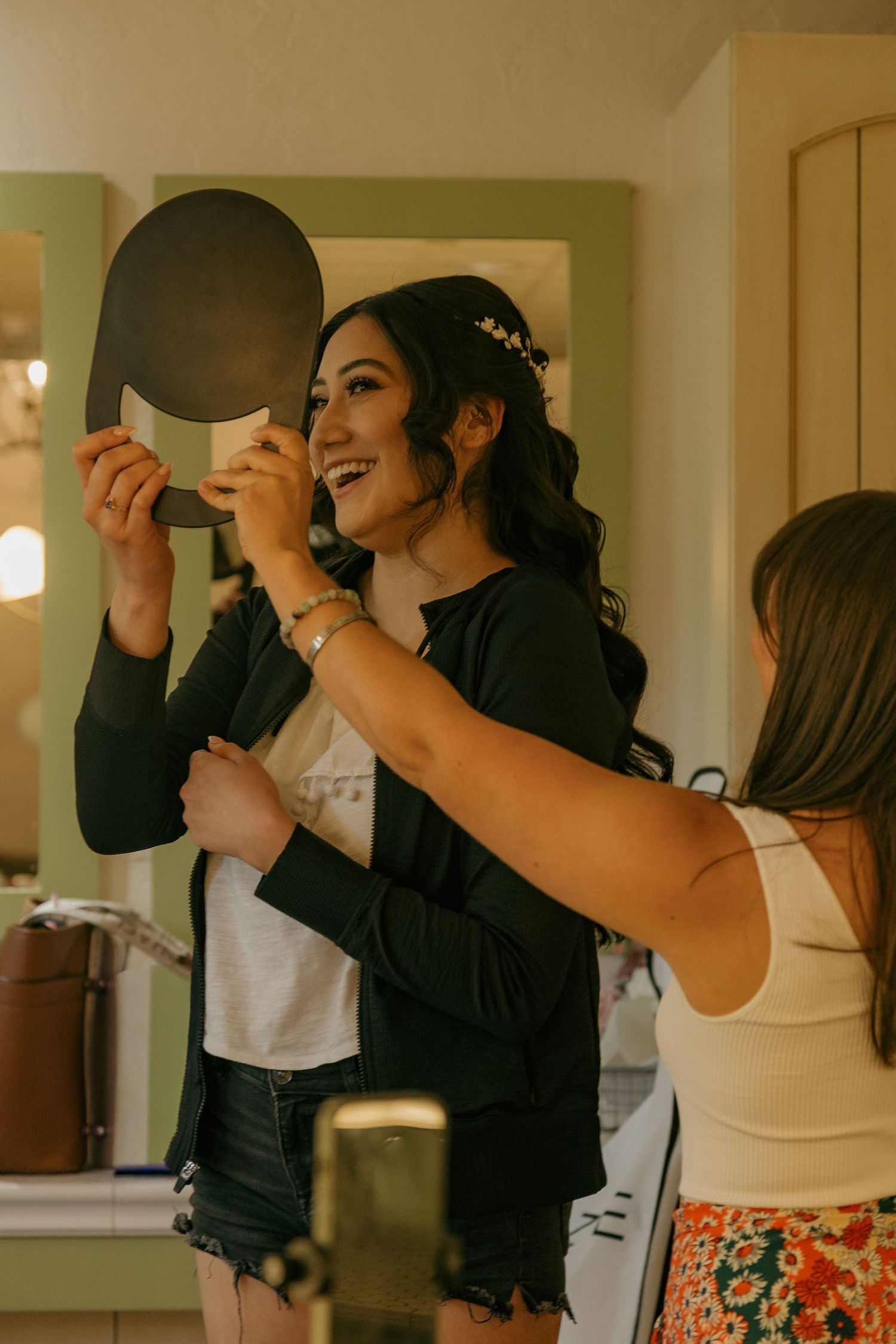 Smiling bride holding a hand mirror admiring her floral hair crown during wedding day preparations.