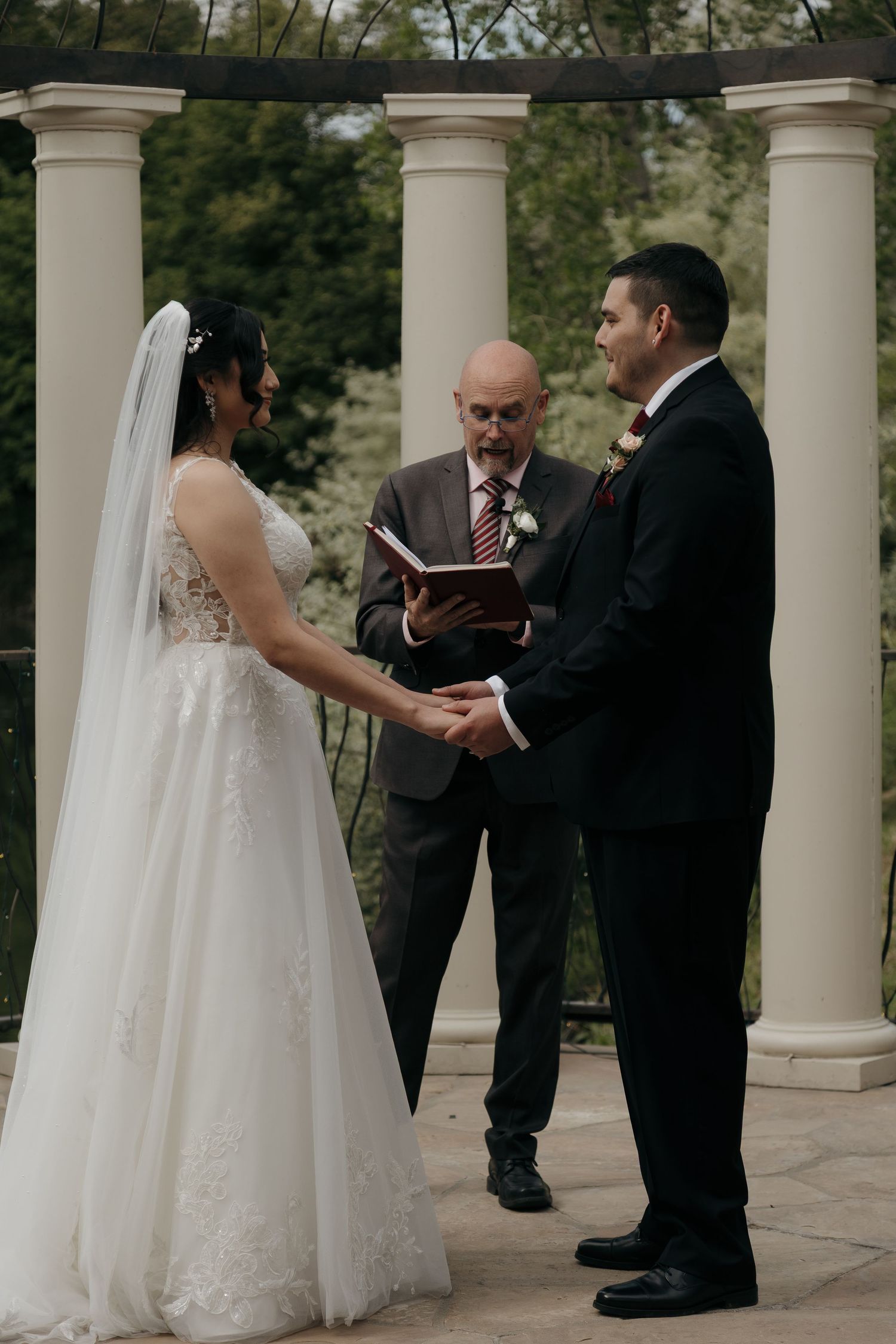 Bride and groom hold hands during wedding vows with officiant reading from book under white columns.