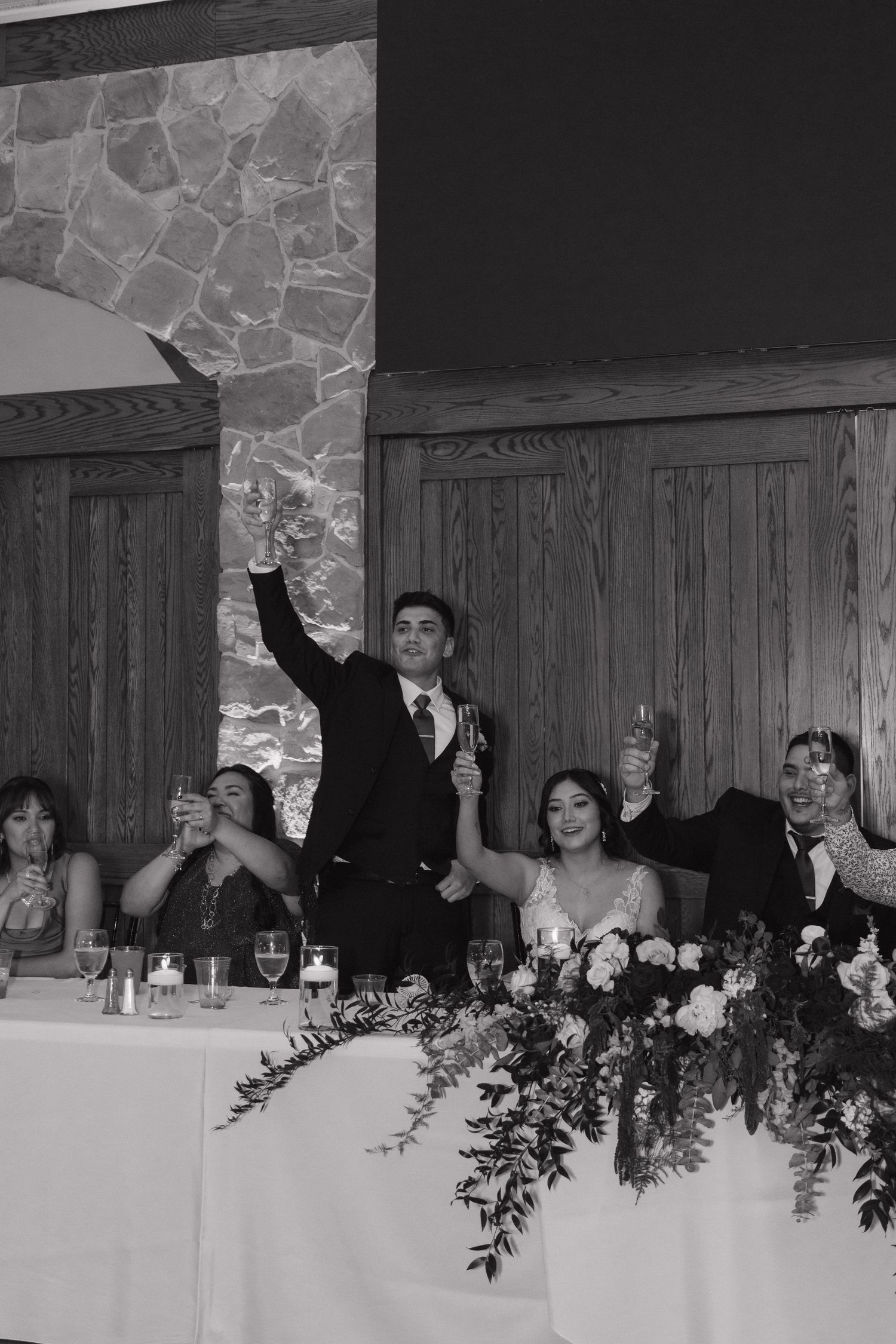 Groom raises a champagne glass in a toast at the wedding head table in black and white.