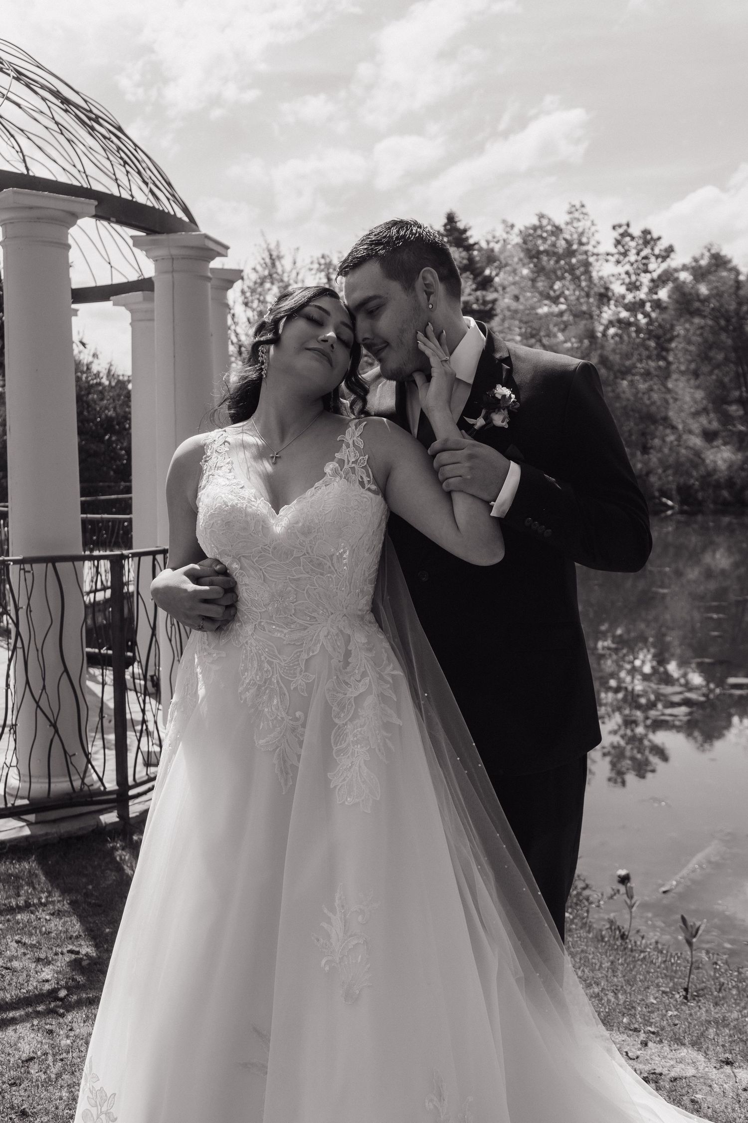 Bride and groom embracing near a gazebo by a reflective pond in black and white wedding photo.